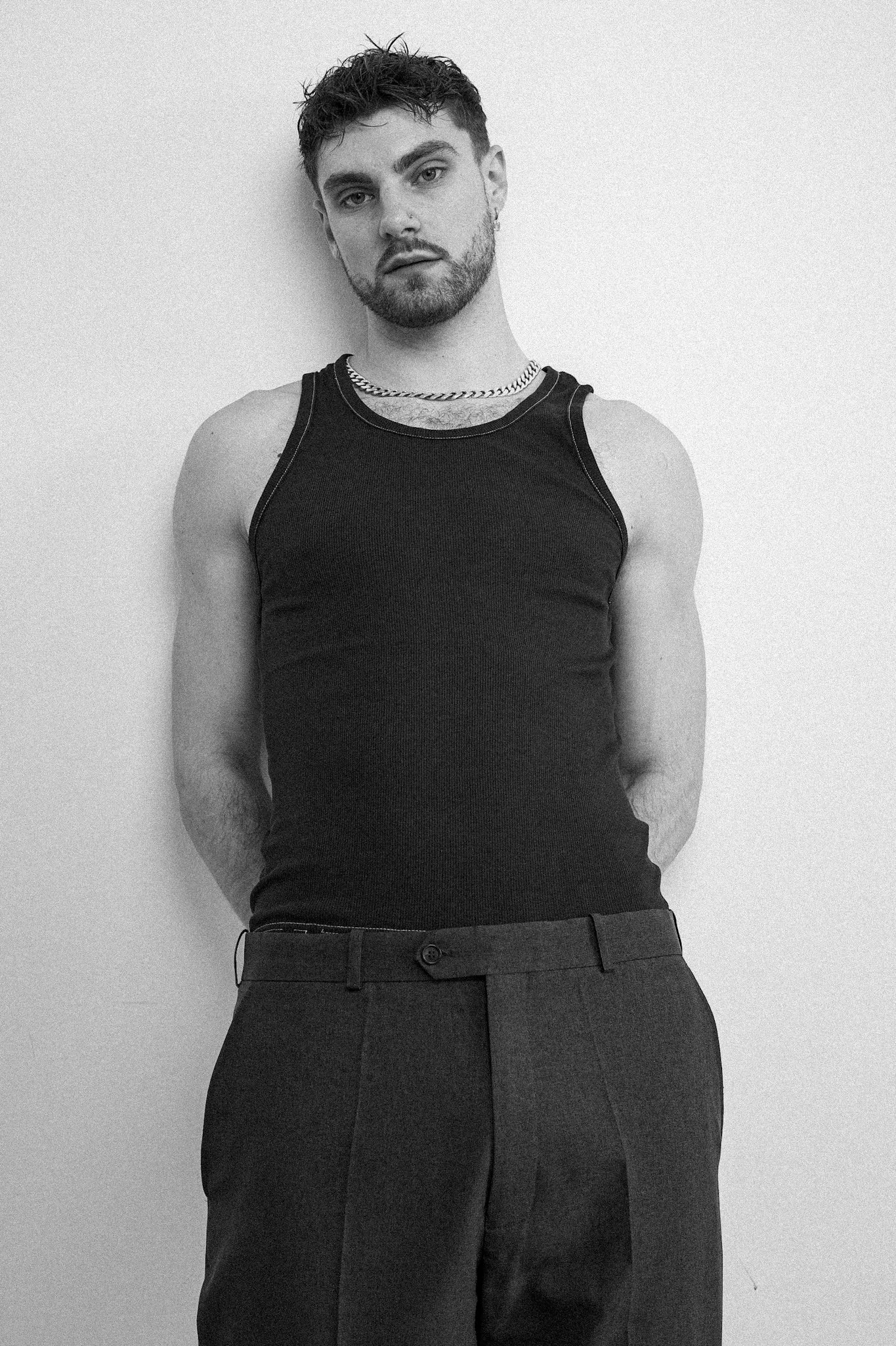 Black and white portrait of a man standing against a plain wall, wearing a dark tank top and chain necklace.