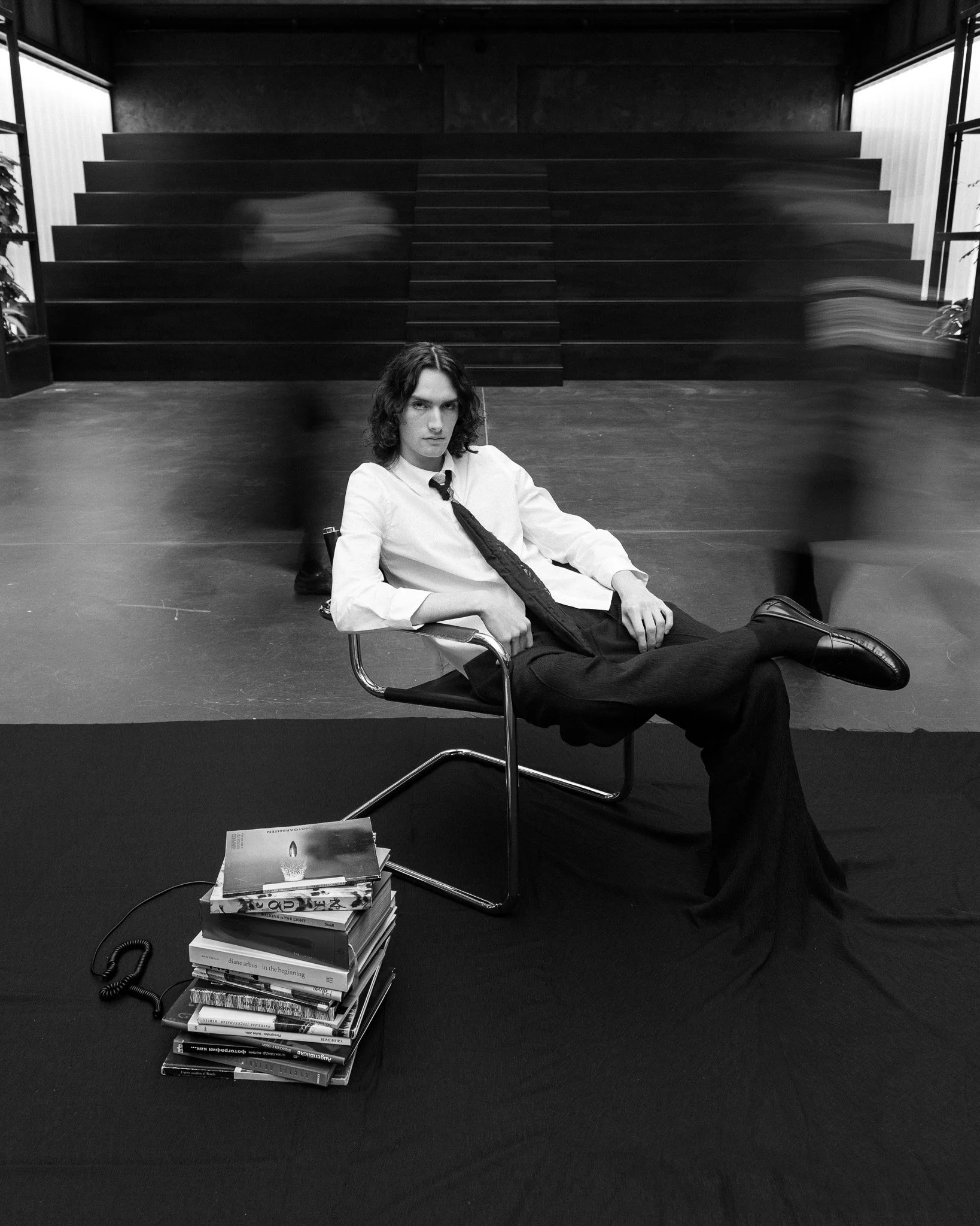 Black and white portrait of a young man sitting in a chair, with blurred figures moving in the background.