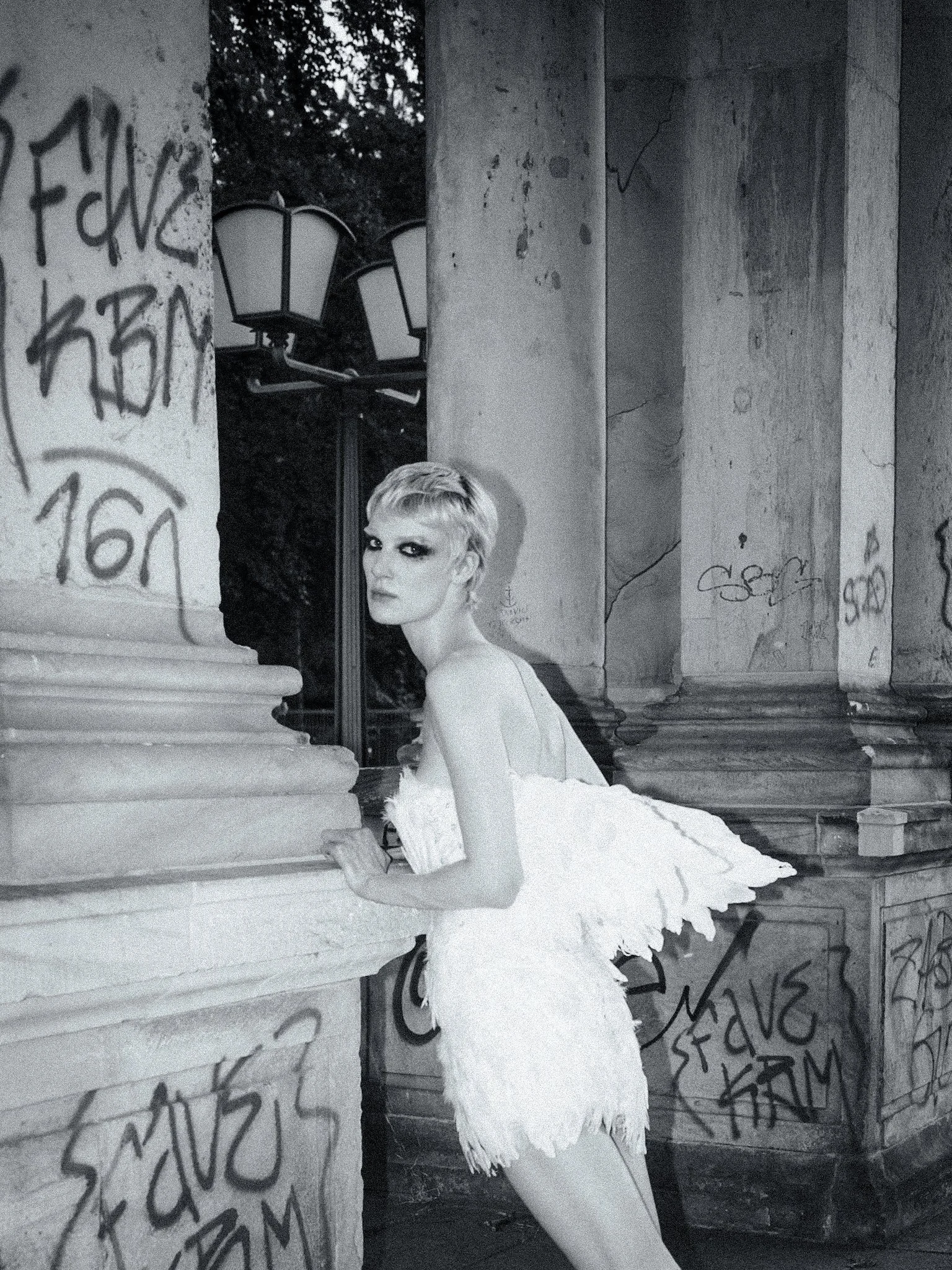Black and white portrait of a woman wearing a feathered dress with wing-like details, standing among graffiti-covered columns in Berlin.
