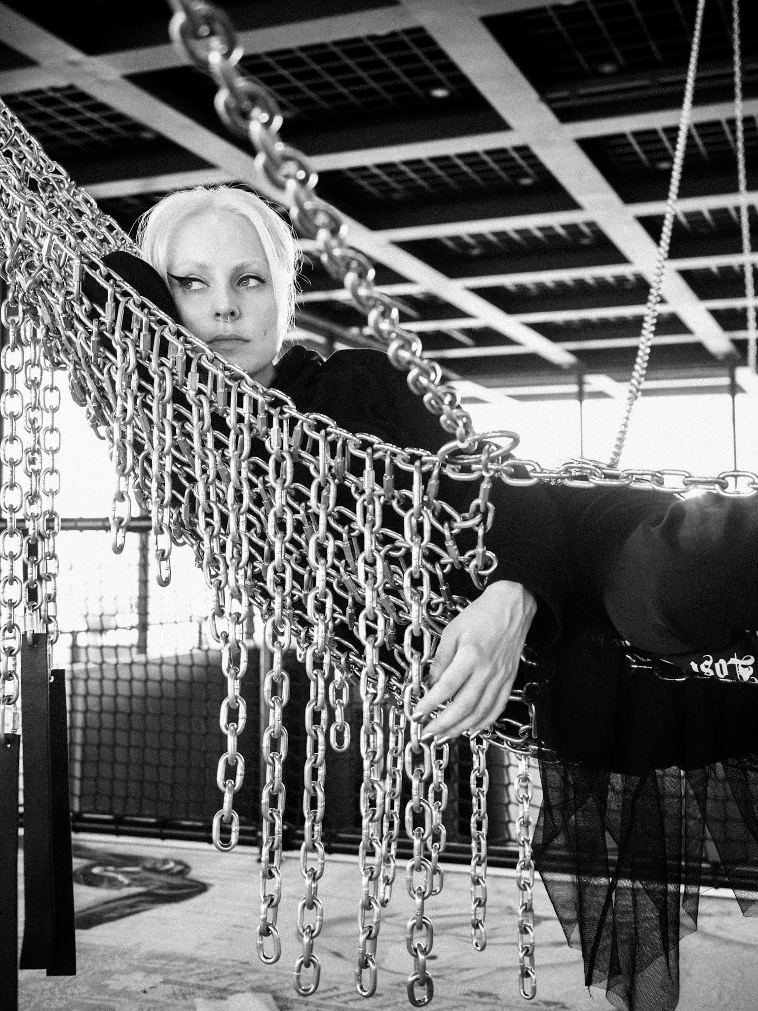 A woman with platinum blonde hair and dark clothing sits on a swing made of metal chains, inside an indoor space with a grid-like ceiling and a mesh fence in the background. The image is in black and white.