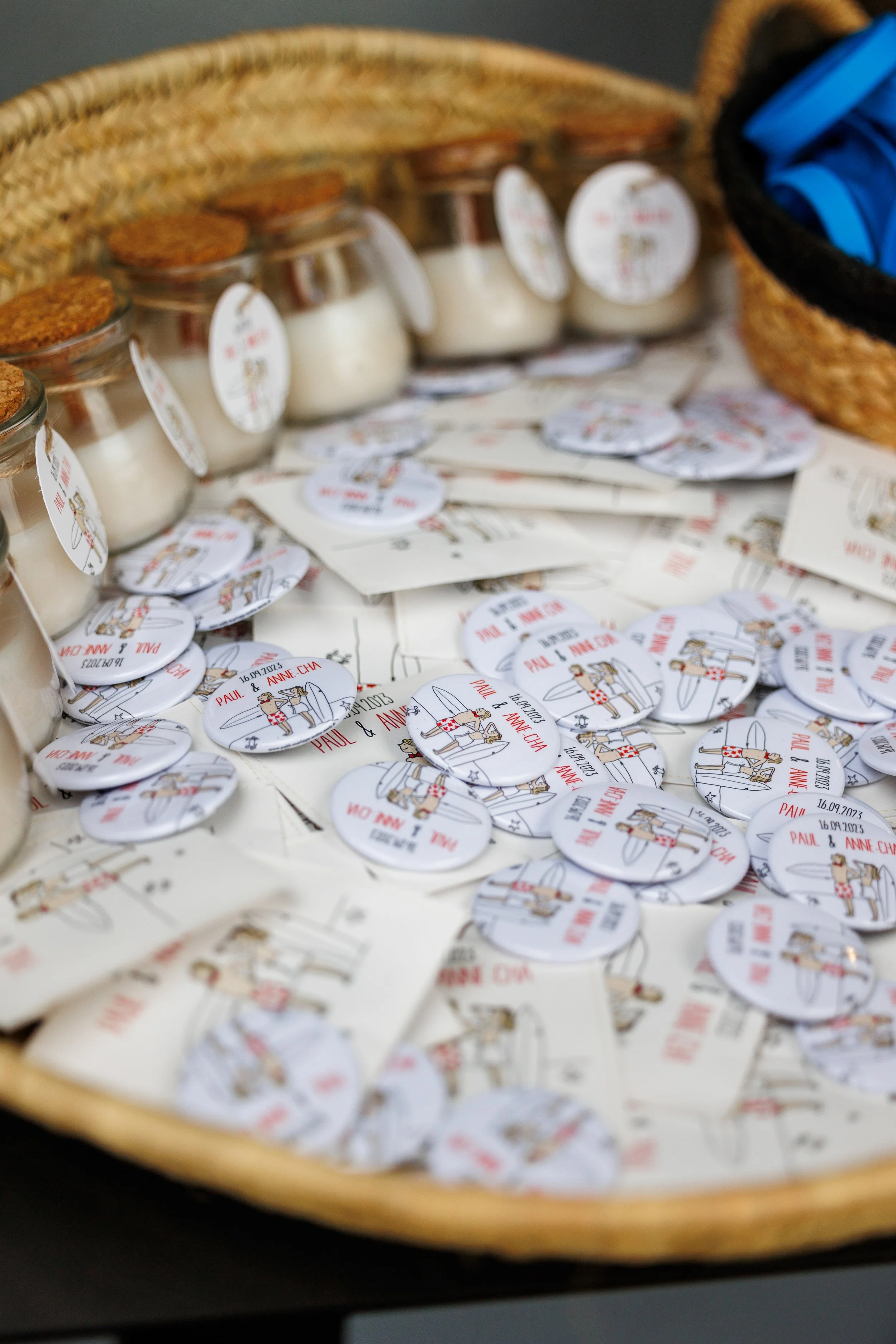Petite jarre blanche avec un bouchon en liège dans un panier en osier, entourée de plusieurs étiquettes rondes avec des dessins de deux personnes en tenue de mariage, portant un texte en français qui semble être une invitation ou un souvenir de maria