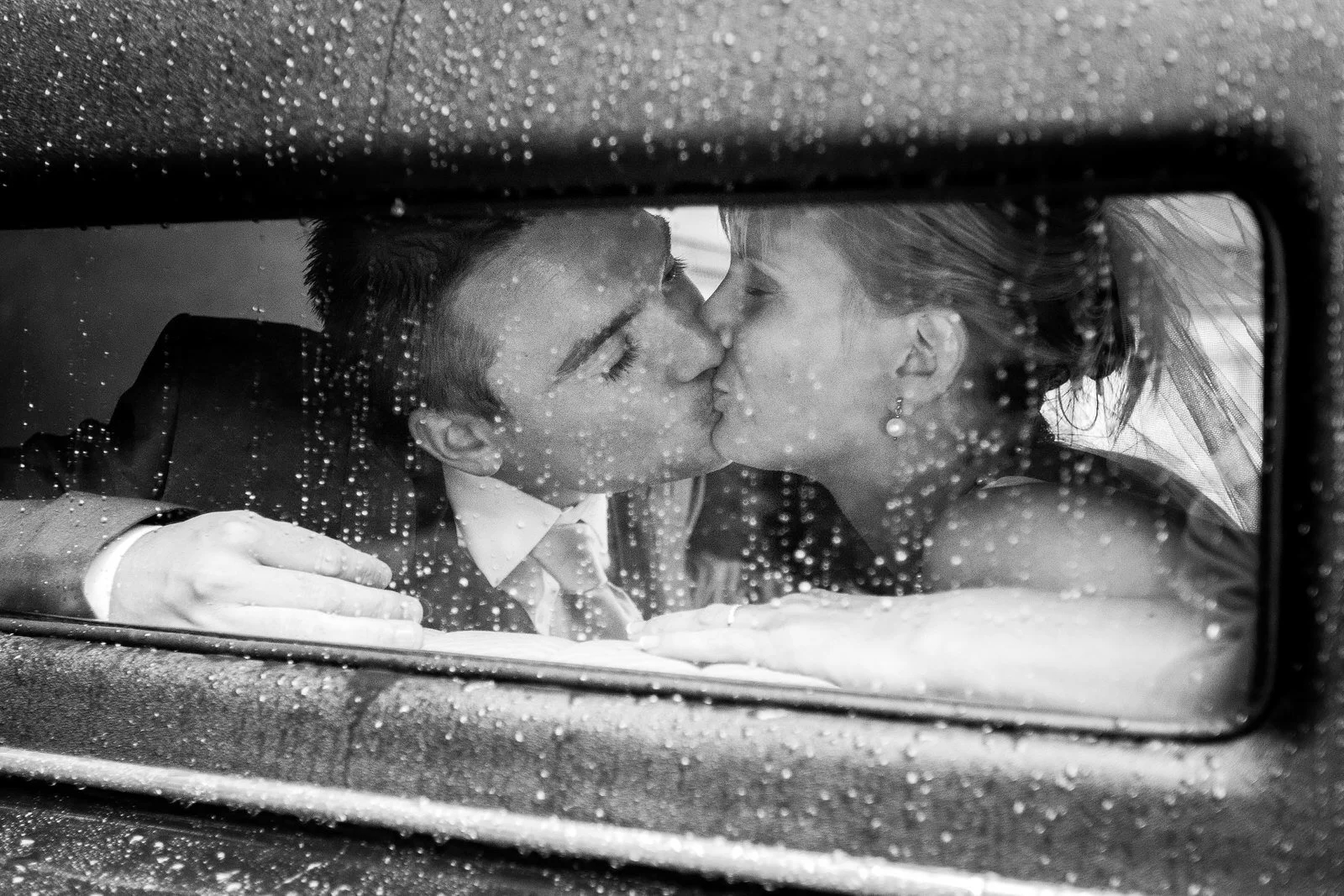 Un couple s'embrasse tendrement à travers une vitre mouillée, en noir et blanc.