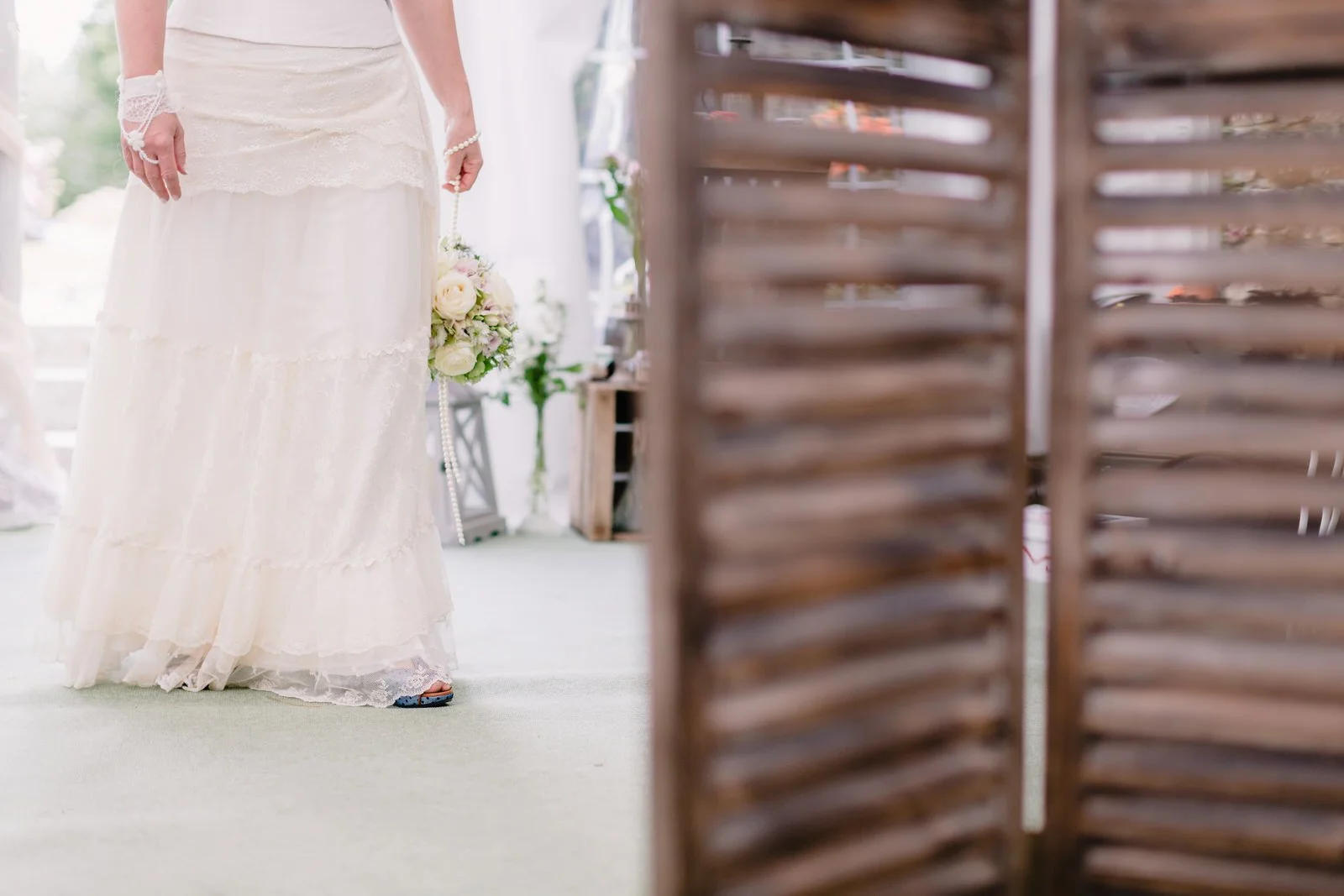 Une femme en robe de mariée blanche tient un bouquet de fleurs dans ses mains. La photo est prise en intérieur avec un écran en bois à droite, et la personne est partiellement visible.