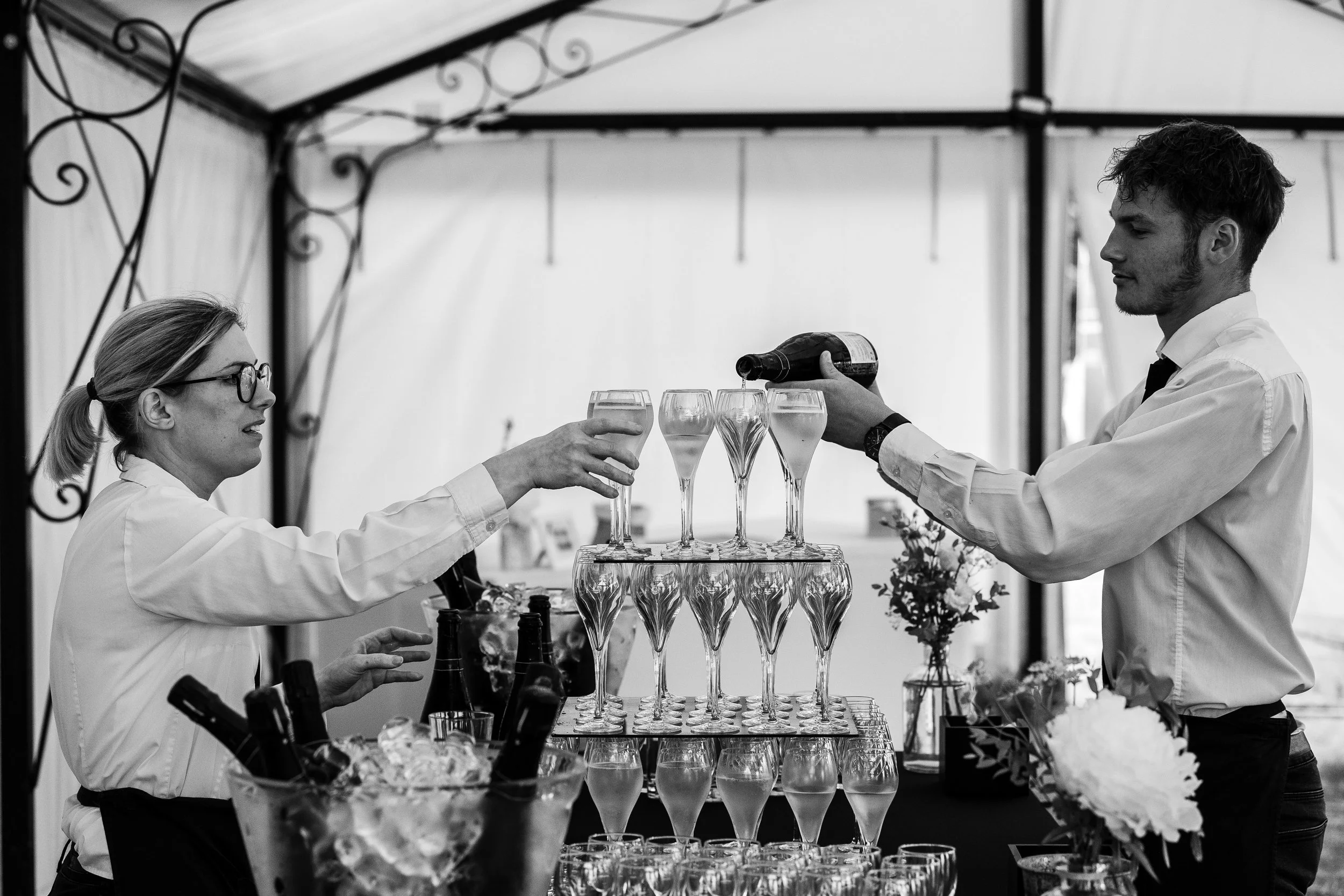 Deux serveurs, un homme et une femme, servent des verres de champagne lors d'une réception, avec une pyramide de verres en arrière-plan.