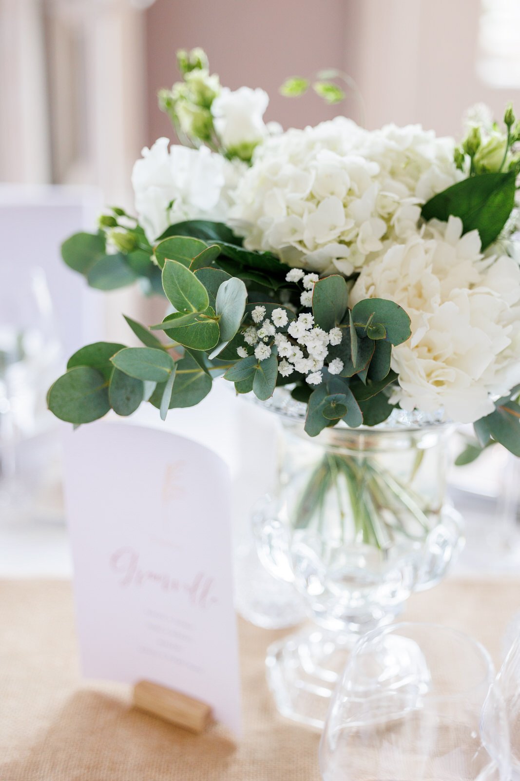 Bouquet de fleurs blanches dans un vase en cristal, sur une table. En arrière-plan, une carte de nom et une petite tablette en bois.