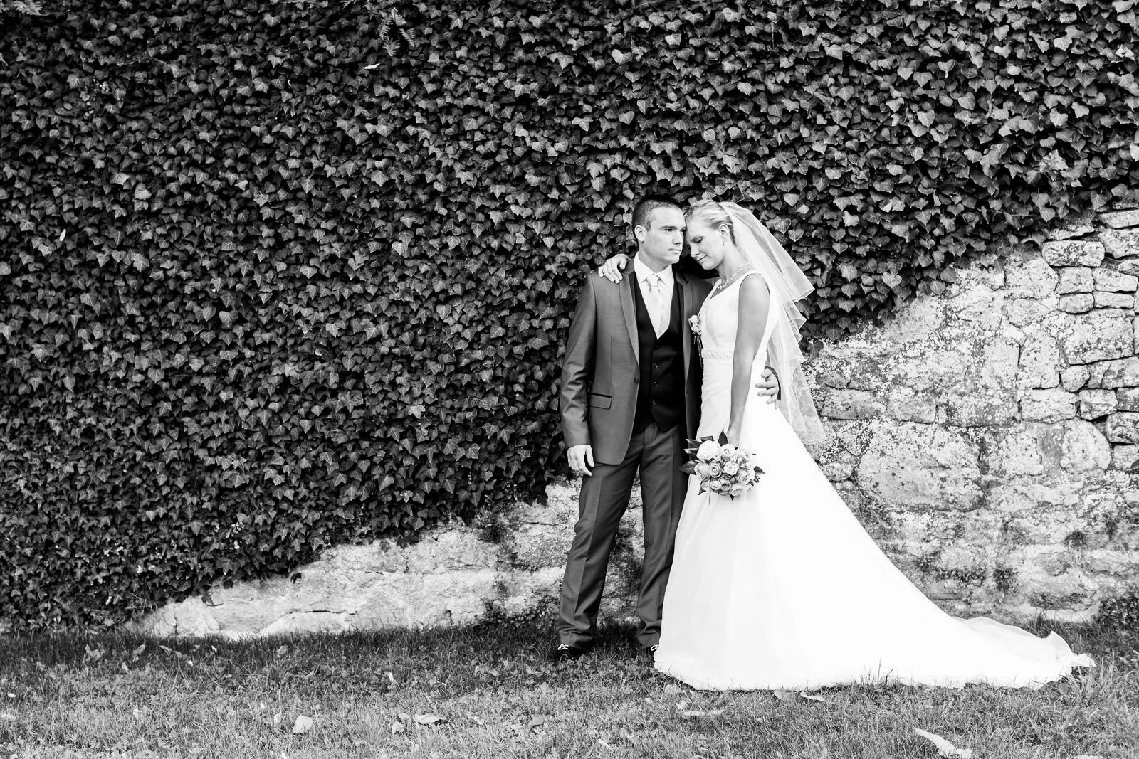 Photo en noir et blanc d'un couple de mariés posant contre un mur de pierres et de feuilles. La mariée porte une robe de mariage et tient un bouquet, tandis que le marié porte un costume.