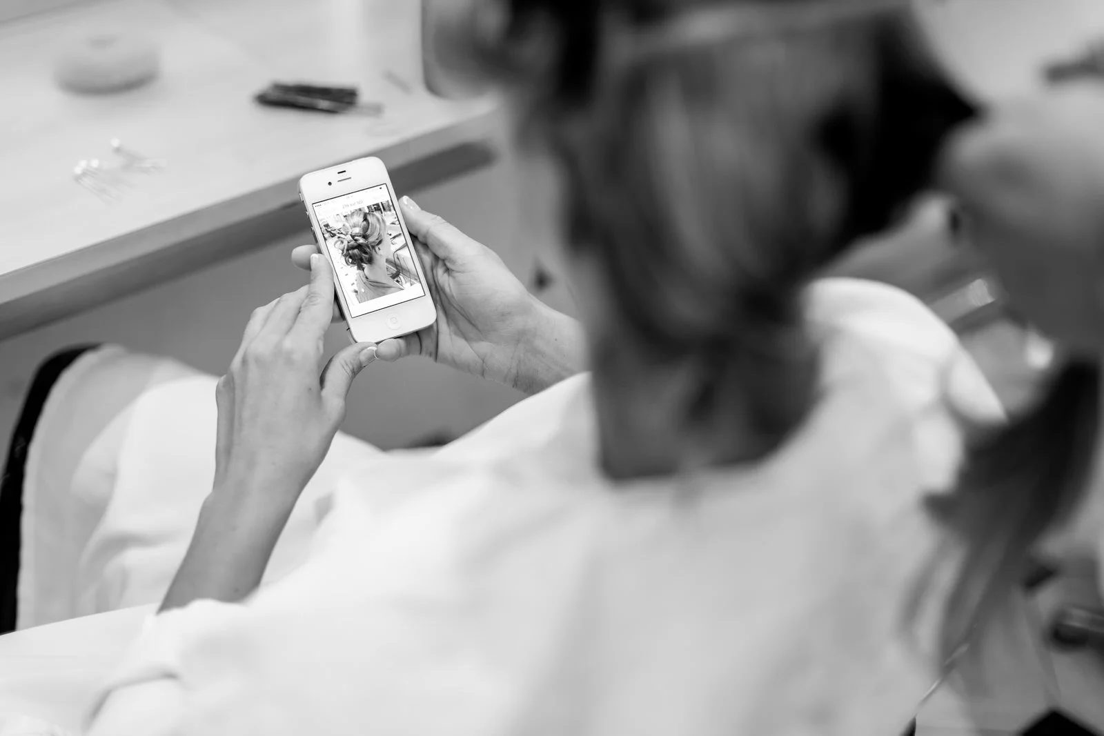 Une personne regarde une photo d'elle-même sur un téléphone, où elle a des cheveux coiffés de manière sophistiquée, dans une pièce avec une table où sont posés des objets divers.