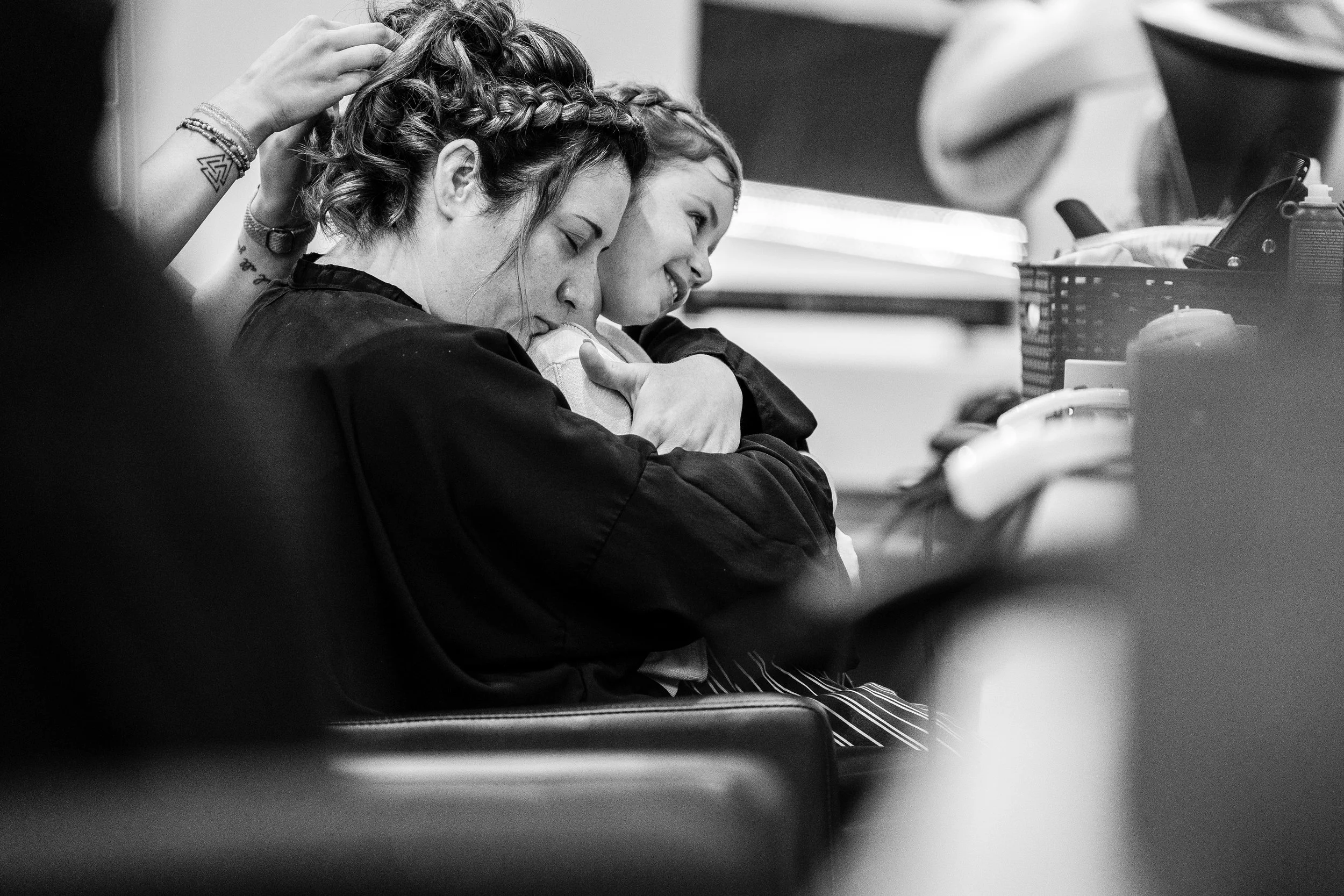Deux jeunes femmes assises sur un banc, l'une d'elles en train de peigner l'autre, dans un environnement intérieur. La scène est en noir et blanc.