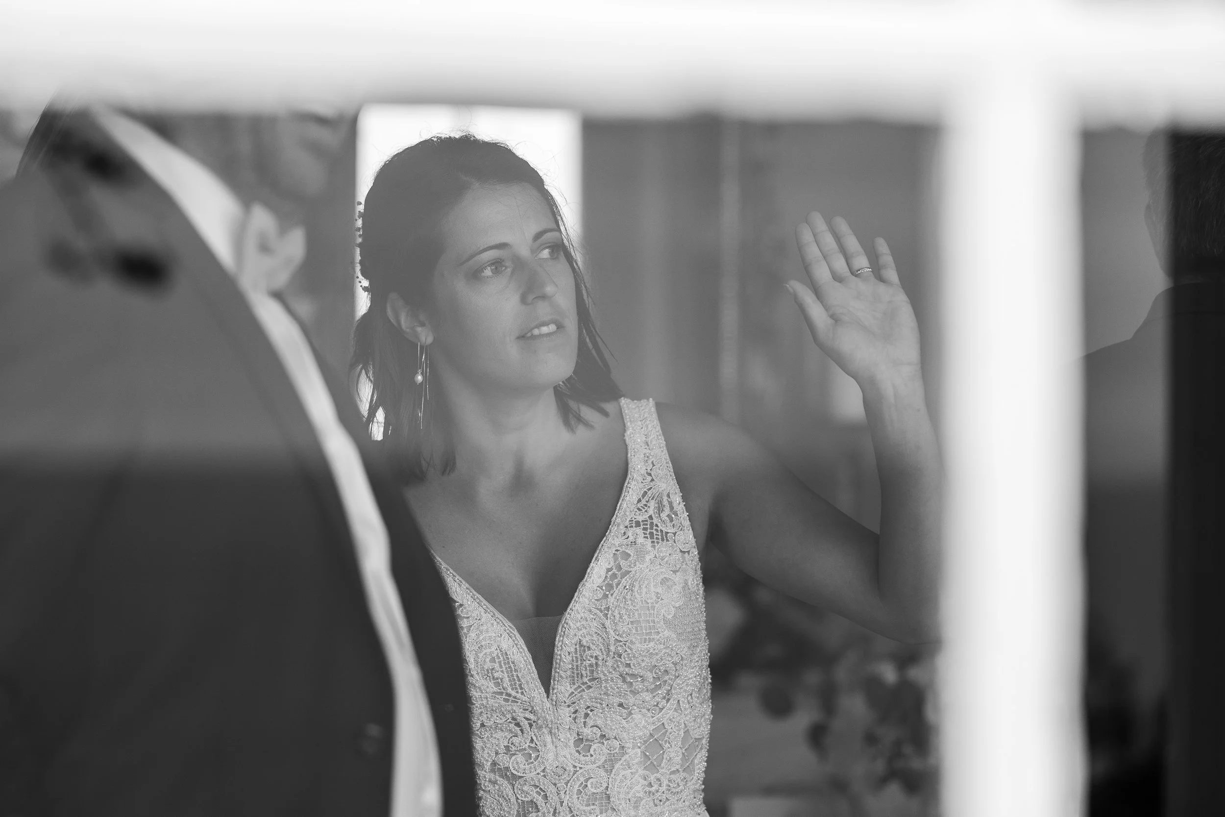 Une femme en robe de mariage parle à un homme lors d'une conversation, vue à travers une vitre ou une barrière en verre.