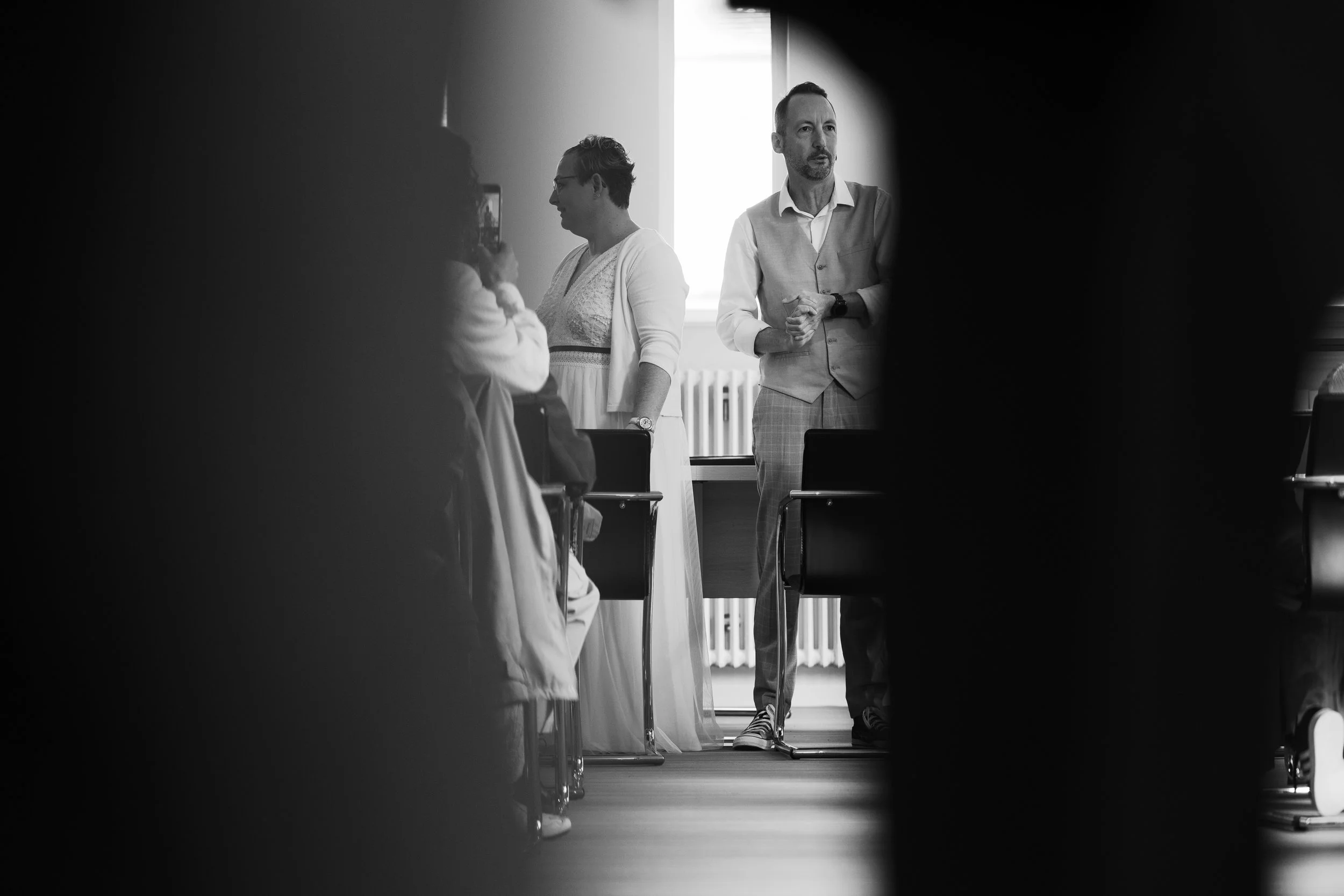 Photo en noir et blanc montrant un homme debout, portant une veste en tissus, parlant à un groupe de personnes en costume. La scène semble se passer dans une salle de réunion ou un auditorium, avec un fond lumineux venant d'une fenêtre.