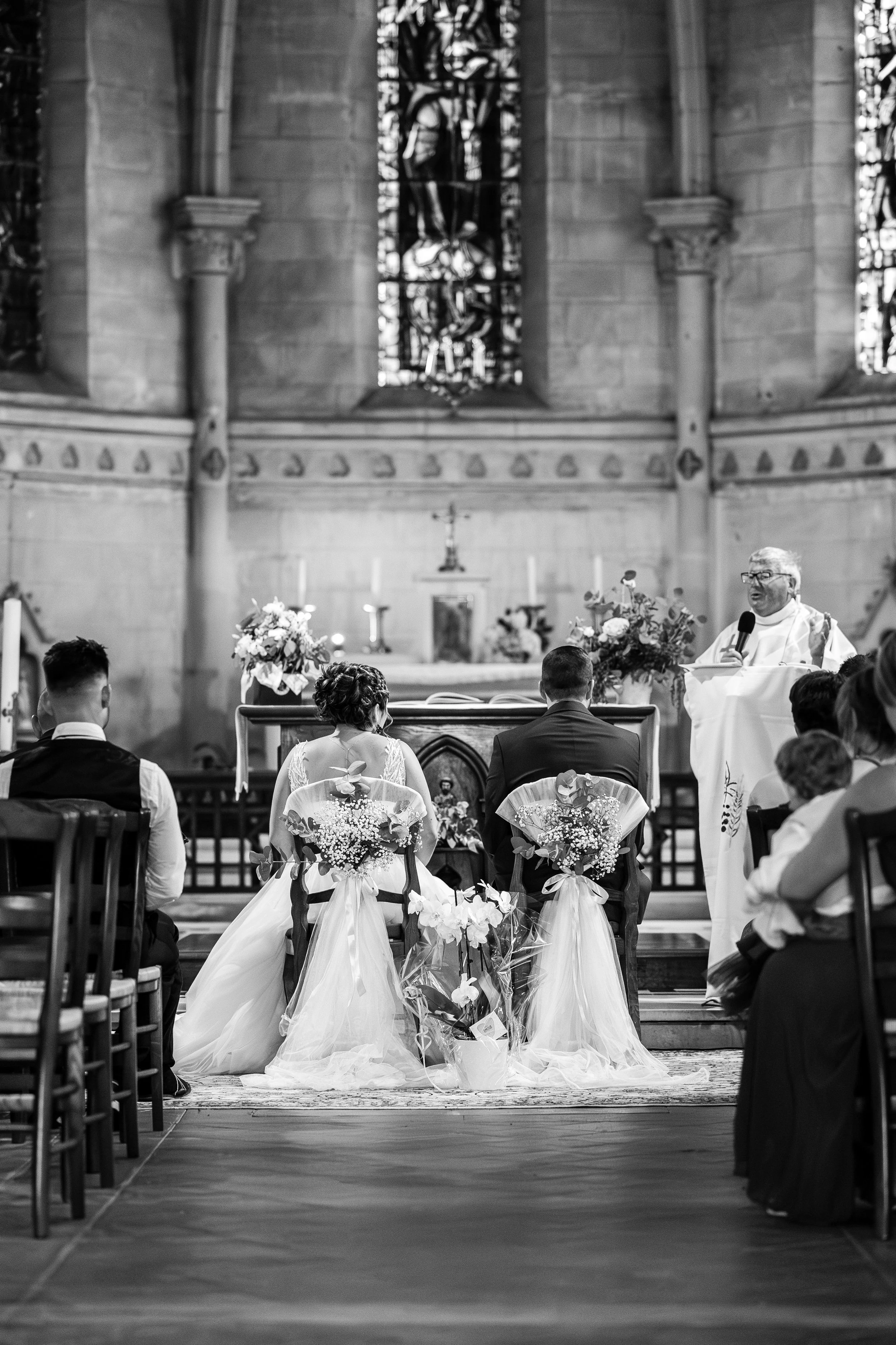 Une cérémonie religieuse de mariage dans une église, avec le prêtre au pupitre, les mariés assis devant, entourés de fleurs et d'invités.