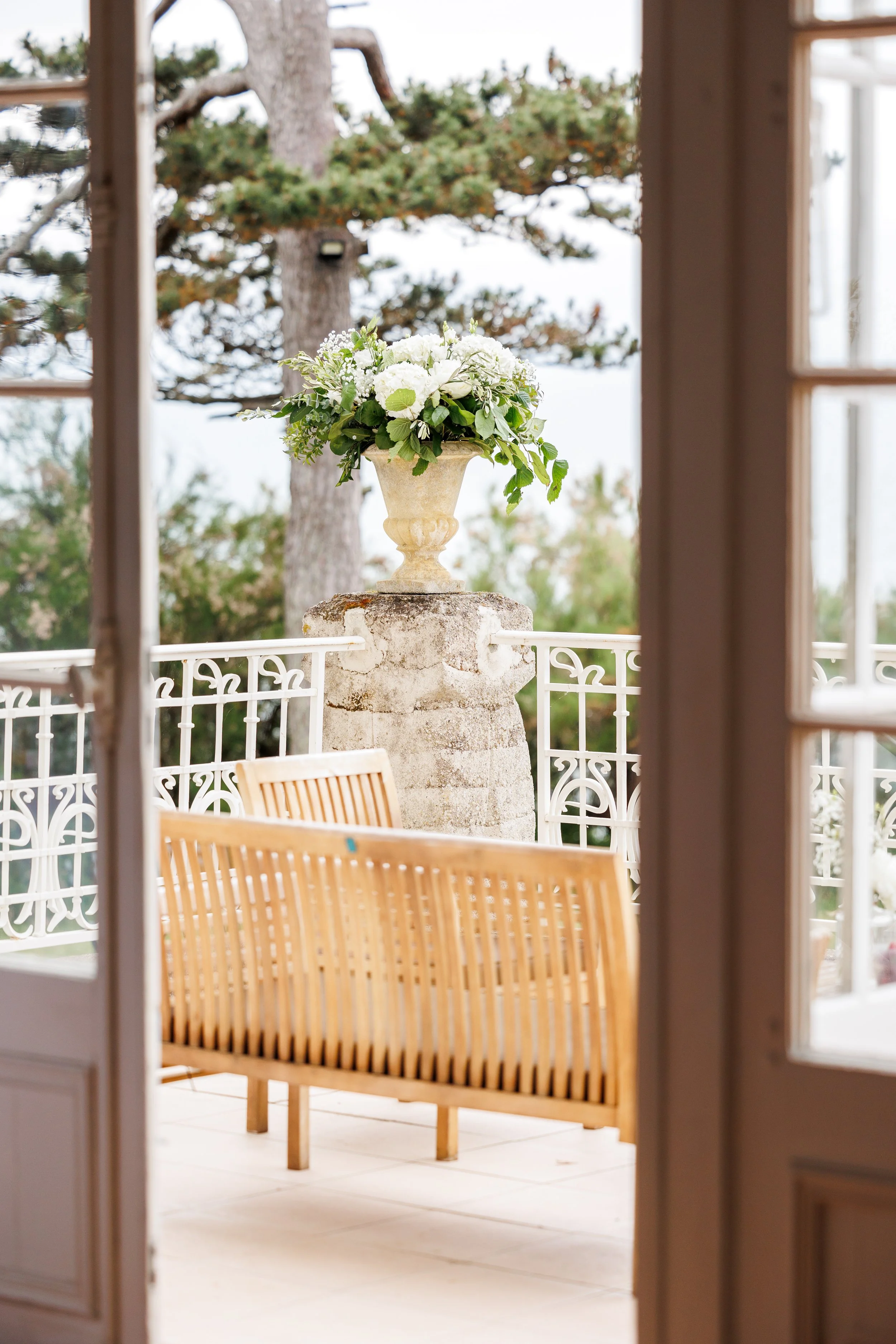 Une vue d'un balcon avec un grand vase de fleurs blanches, entouré d'une rambarde en fer blanc, vue à travers une porte en bois.