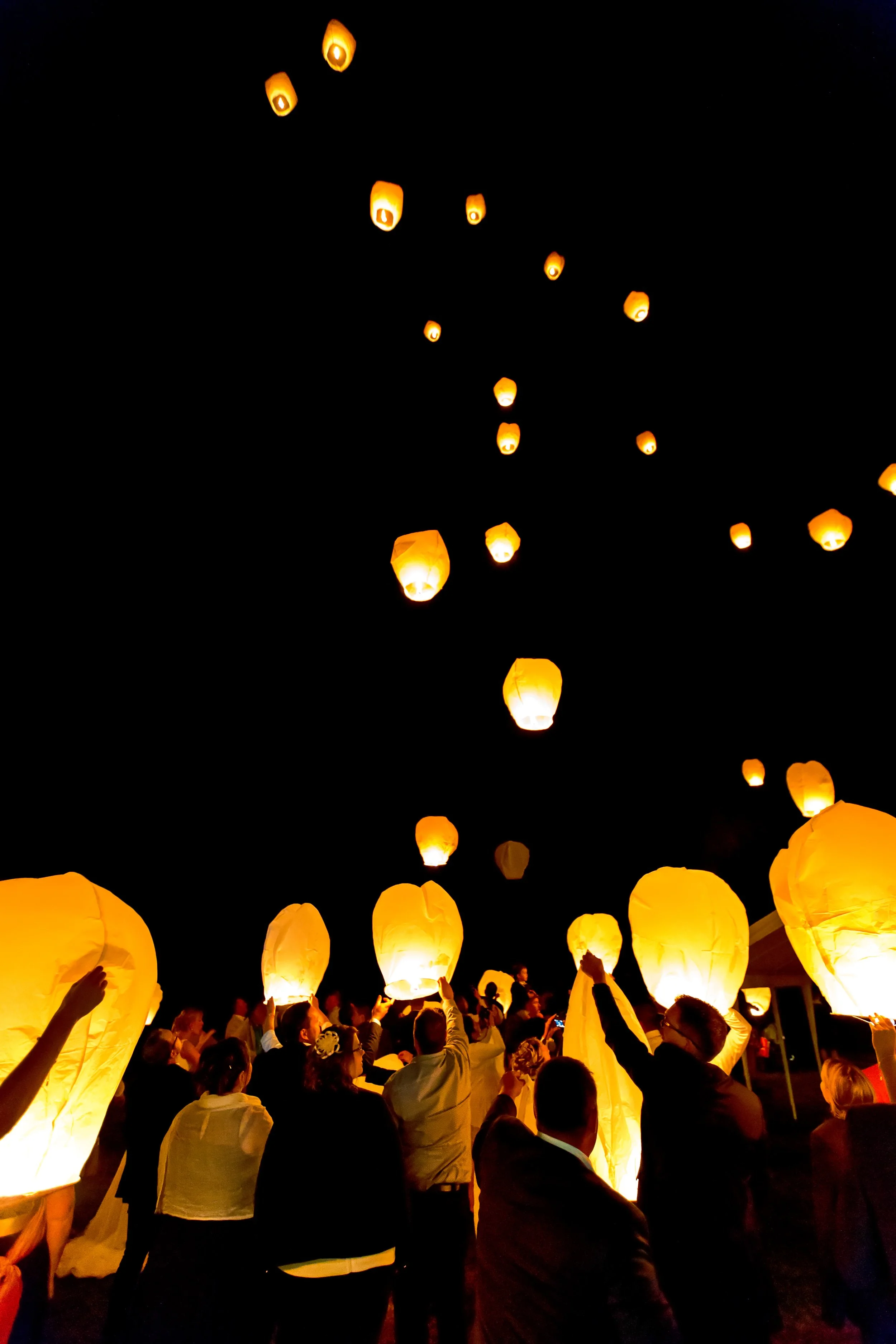 Une foule de personnes lors d'une fête de lanternes en soirée, lançant des lanternes en papier orange dans le ciel noir, créant une scène lumineuse et festive.