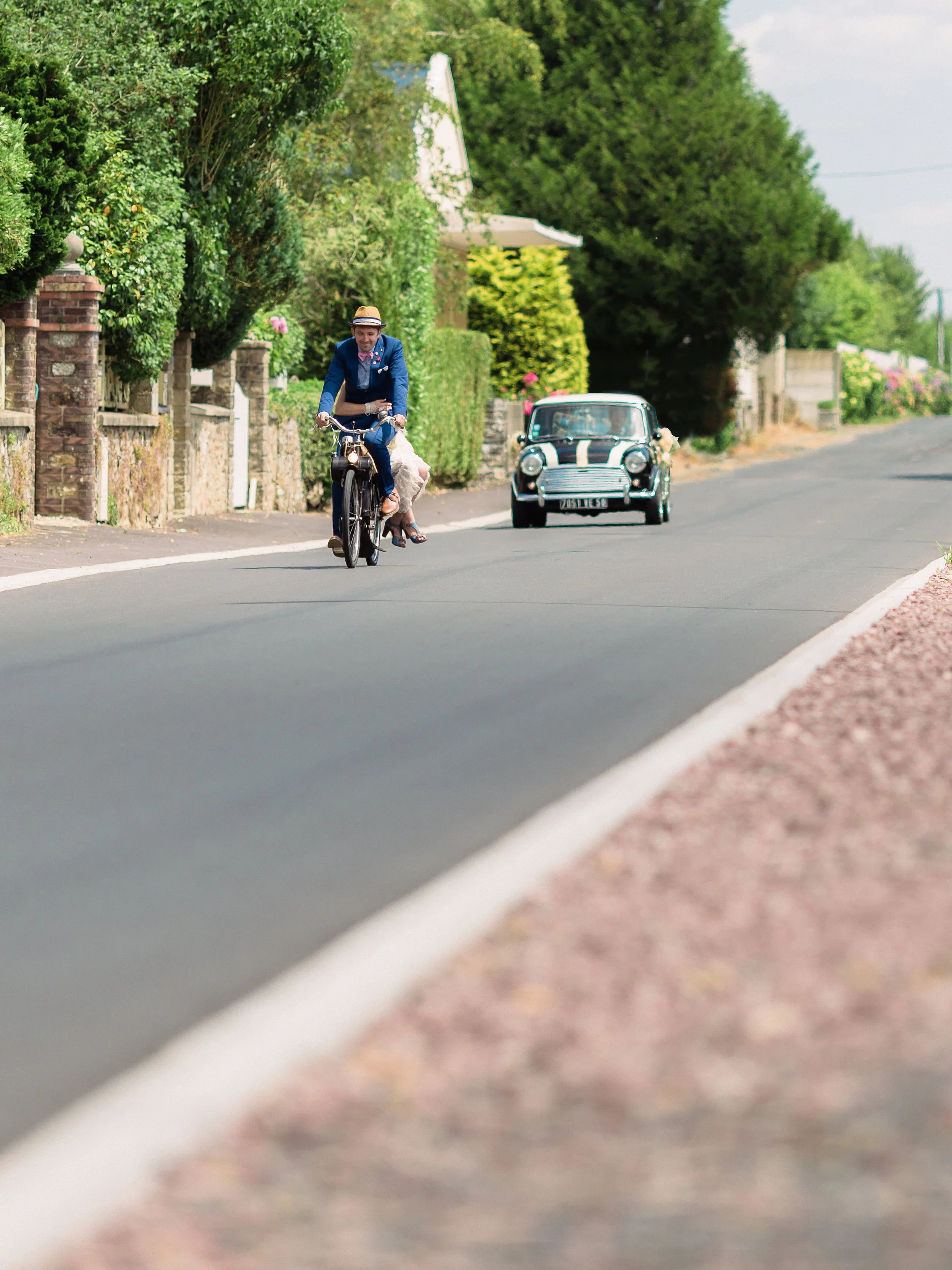 Un homme en costume bleu, portant un chapeau, fait du vélo le long d'une route bordée d'arbres verts, avec une voiture classique derrière lui.
