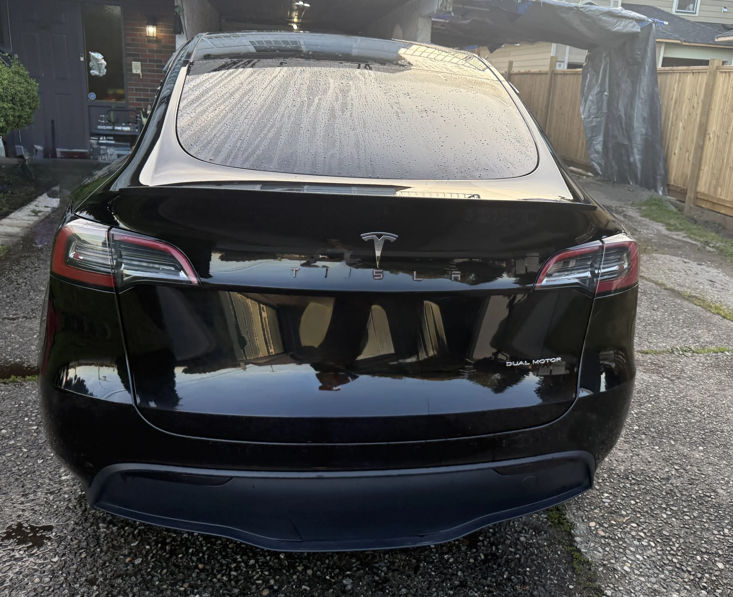 A black Tesla Model S with rain droplets on the rear windshield, parked in a driveway with a wooden fence and house in the background.