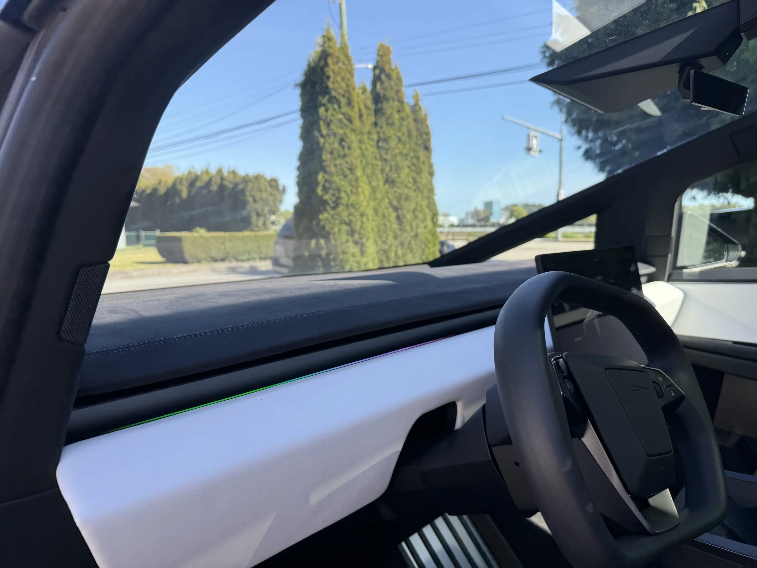 Inside a vehicle showing part of the dashboard, a steering wheel, and the windshield with two tall cypress trees and utility poles visible outside.