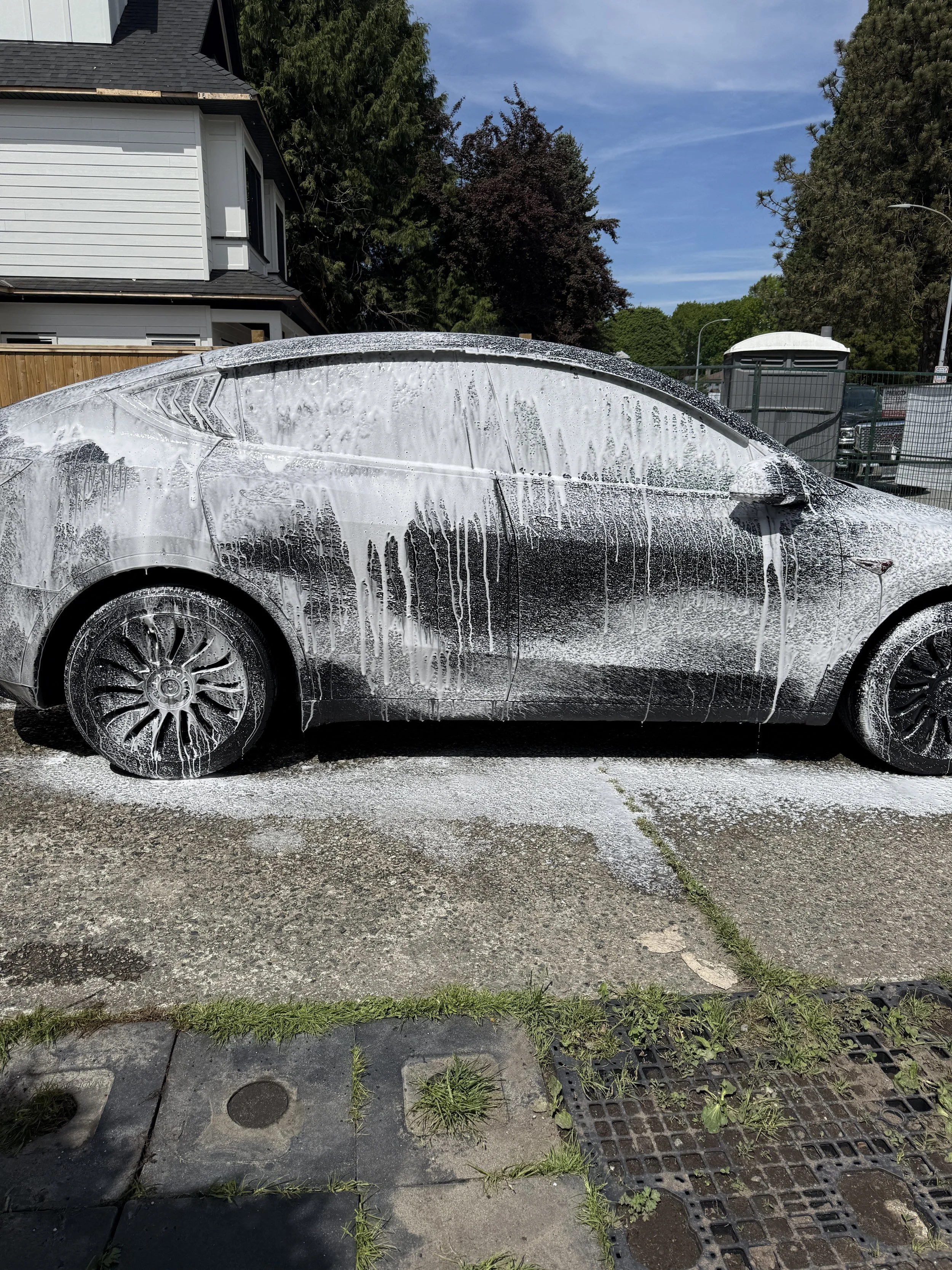 A white car covered in soap foam parked on a concrete driveway with some grass and plants growing through the cracks, residential houses and trees in the background, and a cloudy blue sky overhead.