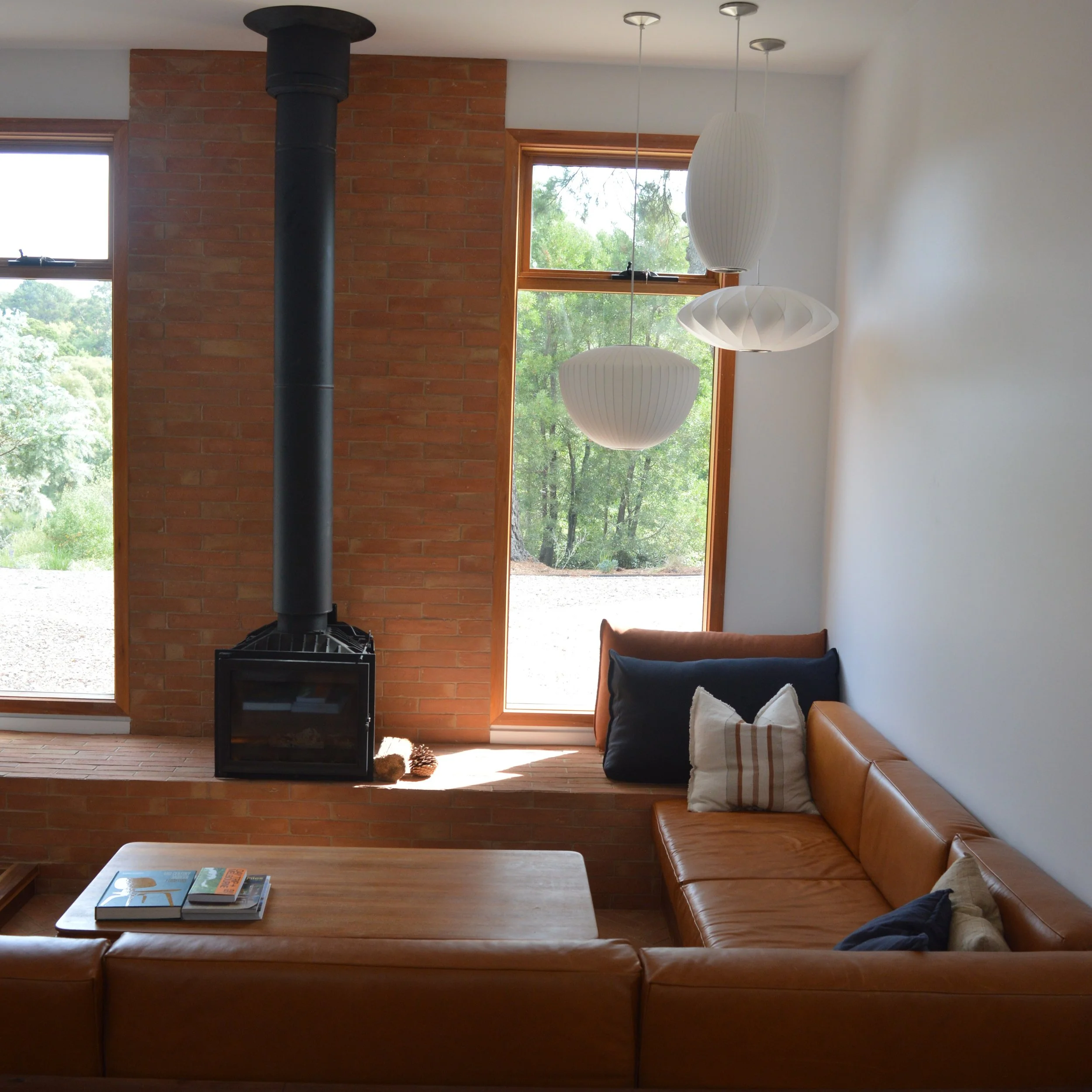 Living room with large windows, a black fireplace with a chimney, a wooden coffee table with books, a brown leather couch, and modern hanging pendant lights.