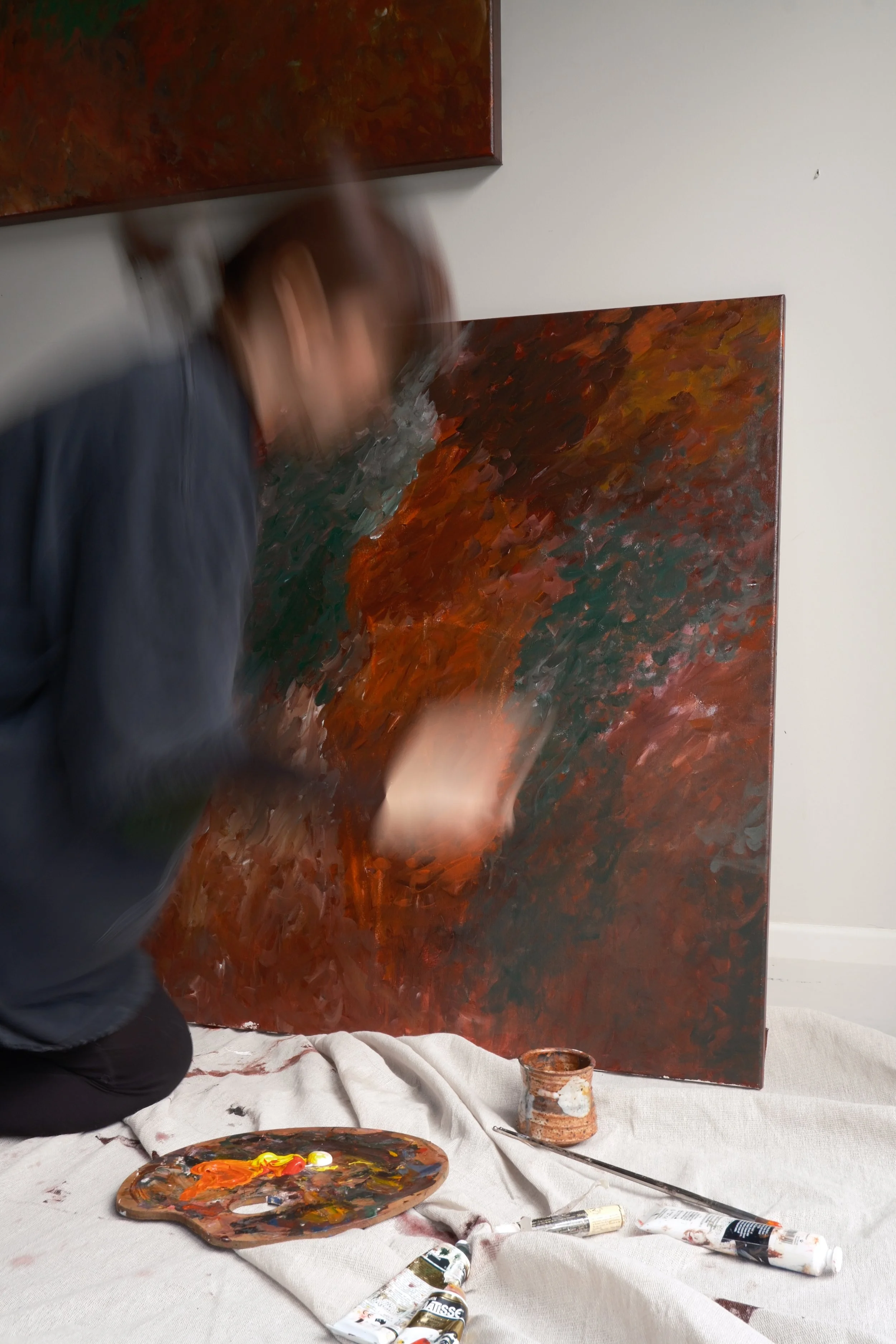 Artist creating an abstract painting with bold, textured brushstrokes in warm colors on a large canvas in a studio. Art supplies, including a painter's palette with mixed colors, tubes of paint, and a palette knife, are scattered on a white cloth cov