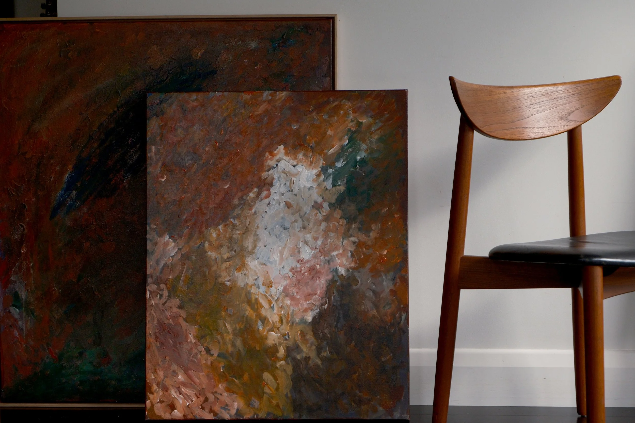 Two abstract paintings with thick, textured brushstrokes in warm earth tones, leaning against a white wall next to a wooden chair with a curved backrest.