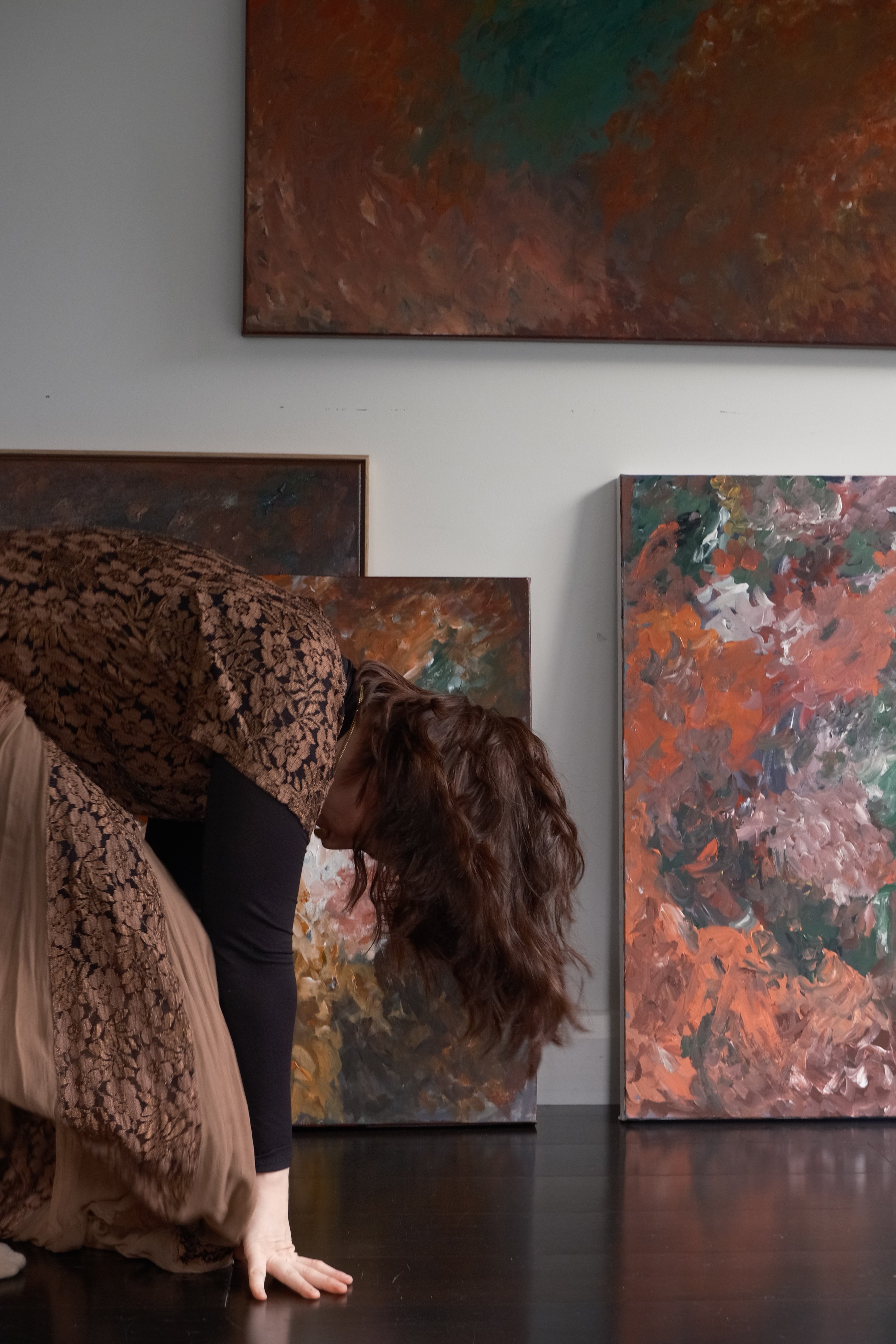 A person with long, curly hair bends forward in front of two large, abstract expressionist paintings hanging on a white wall. The paintings are colorful, with one featuring autumnal colors and the other in darker shades.