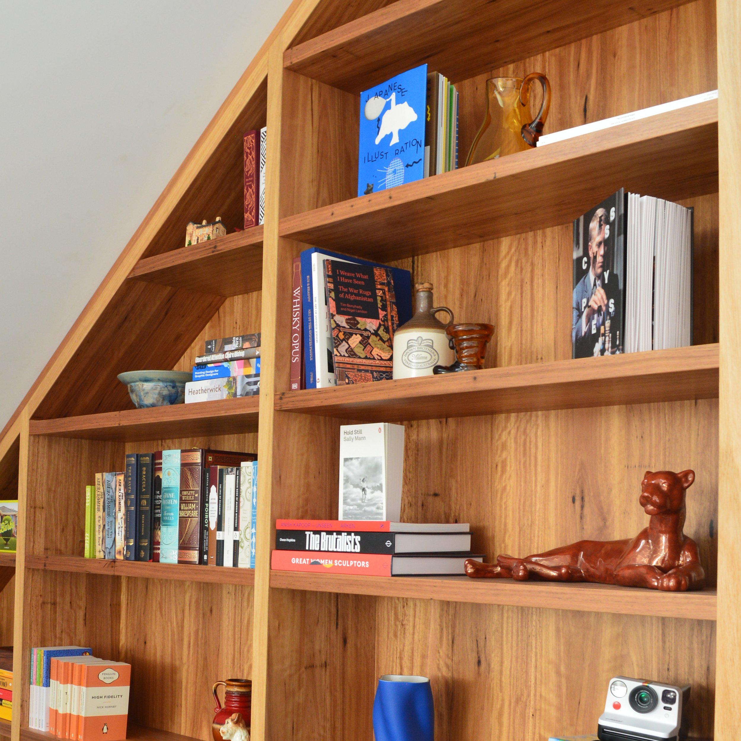 A wooden bookshelf with multiple shelves holding books, vases, a ceramic dog figurine, and a polaroid camera.