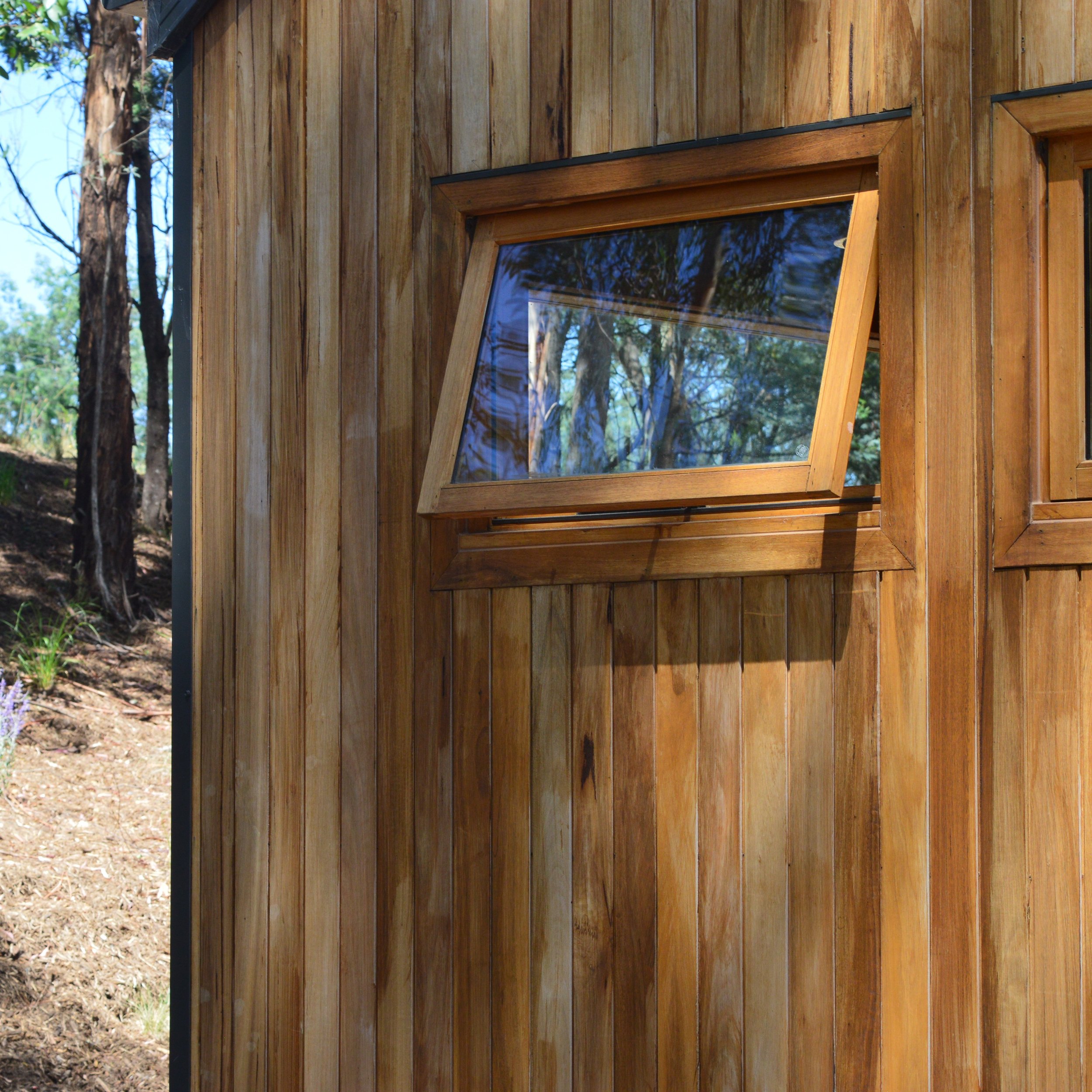 Close-up of a building exterior with wooden vertical planks and a rectangular window, surrounded by outdoor trees and sunlight.