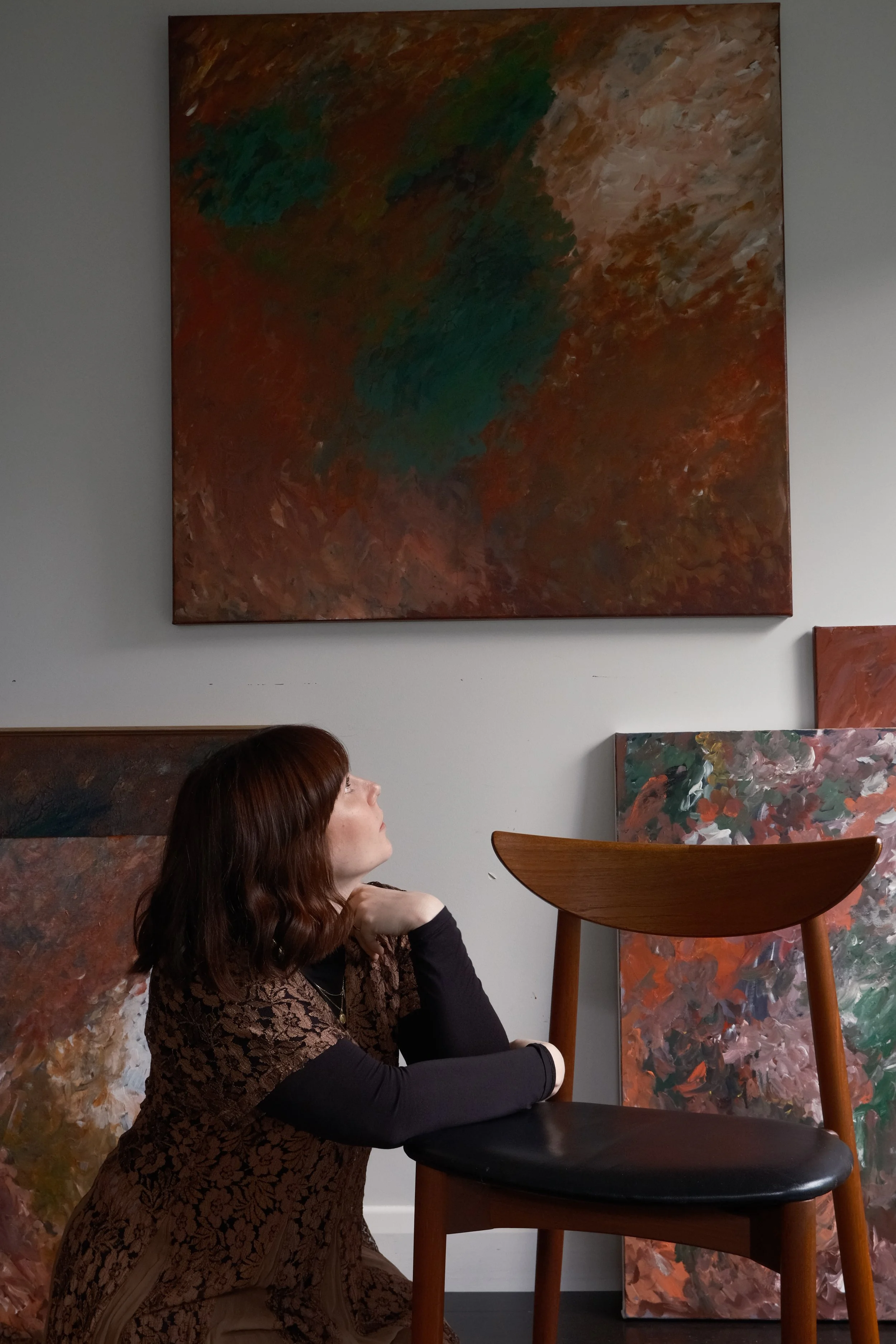 A woman with short  auburn hair, wearing a black long sleeve top and a brown patterned dress, is sitting on the floor in an art gallery, looking thoughtfully at paintings on the wall, with her hand resting near her chin.