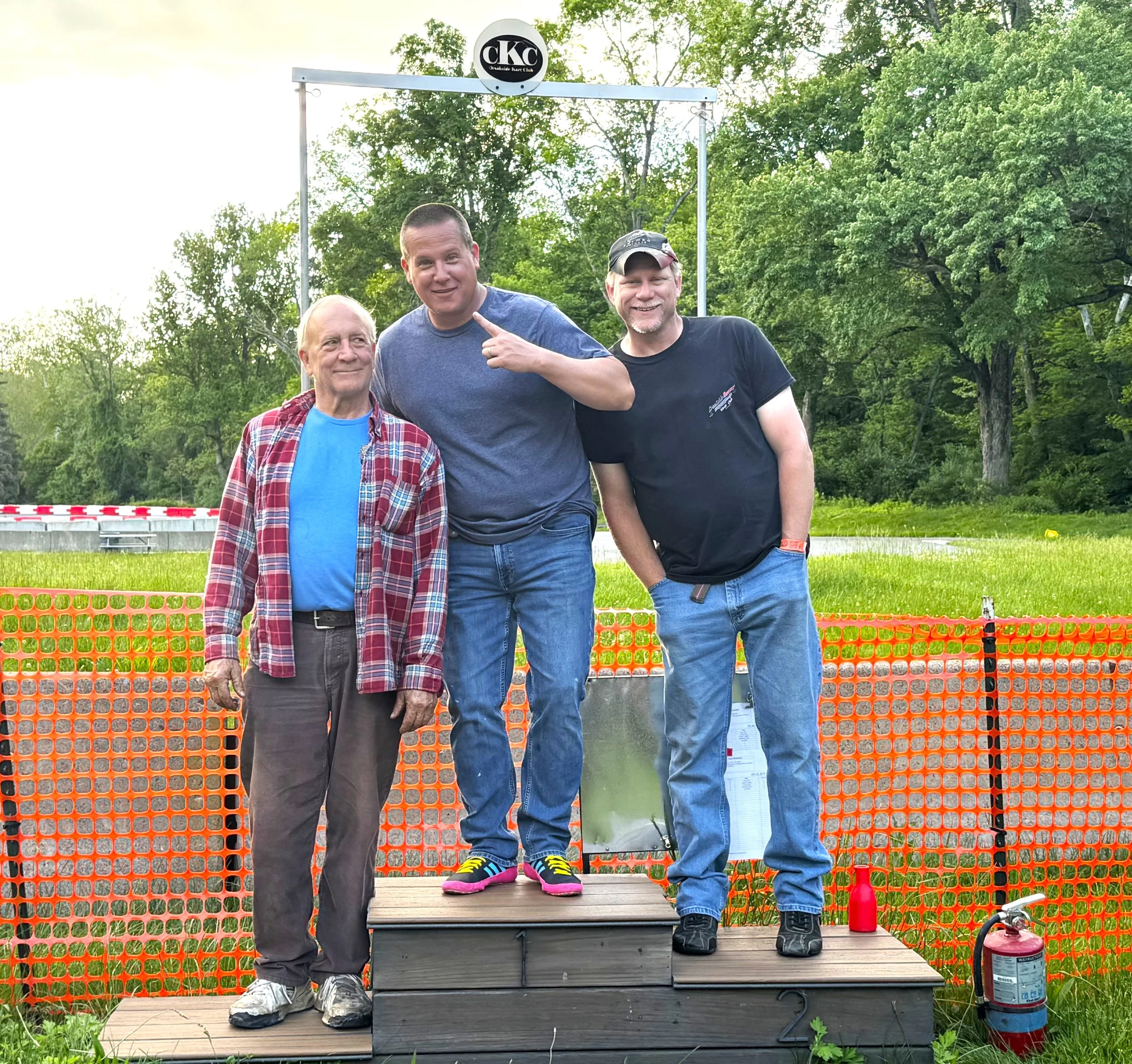 Three men standing on a podium with a woman in a mask and athletic shoes in the middle. They are outdoors, surrounded by trees, with orange safety fencing behind them and a fire extinguisher nearby.