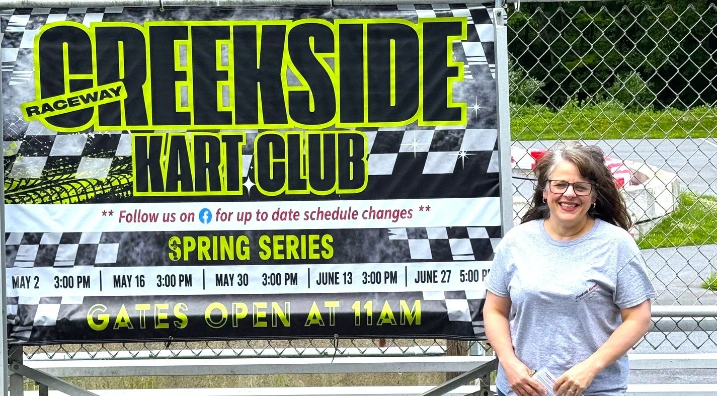 A smiling woman with glasses and long wavy hair standing outdoors next to a large raceway kart club banner. The banner advertises a spring series race event scheduled for May 2, May 16, May 30, June 13, and June 27, with gates opening at 11 am.