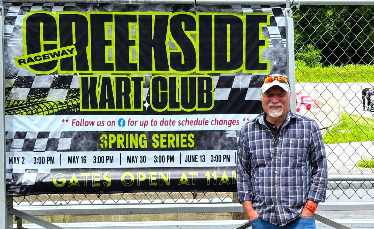 A man wearing a plaid shirt and white cap standing in front of a sign for Hillside Raceway Kart Club, promoting spring racing series with scheduled dates and time, next to a chain-link fence.