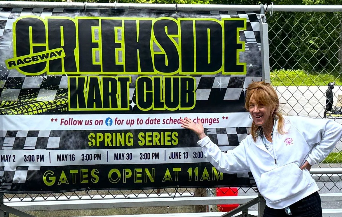 A smiling woman standing next to a sign for the Greeakahs Lakeside Kart Club Racing Track, announcing a spring series event with dates and gate opening time.