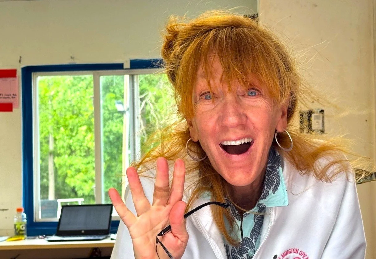 A woman with long, reddish hair, wearing hoop earrings, smiling and waving at the camera inside a room with a window showing green trees outside.