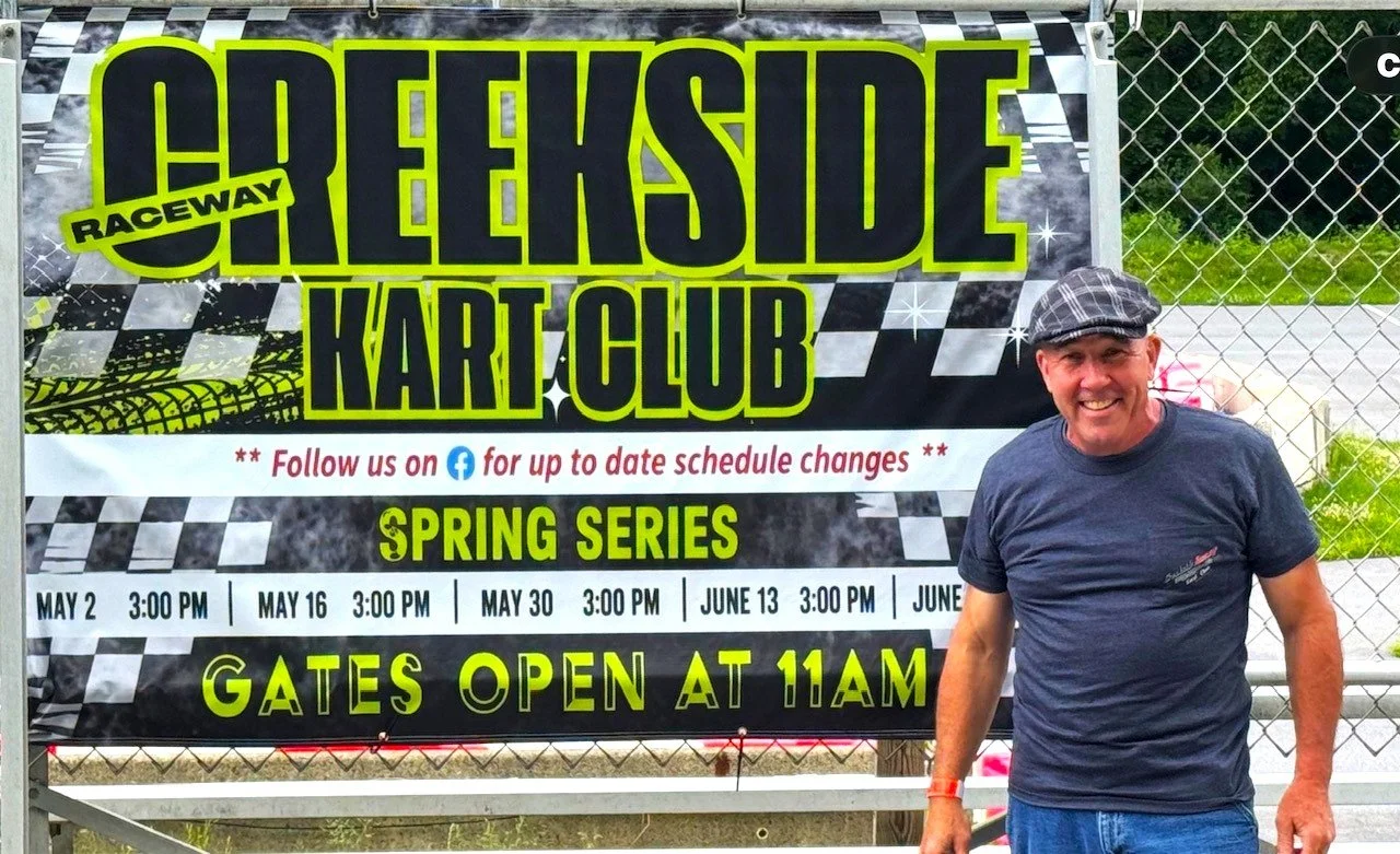 A smiling man wearing a gray flat cap and a dark blue t-shirt standing in front of a sign for the Lakeside Raceway Kart Club spring series. The sign displays dates and times for races and indicates that gates open at 11 a.m.