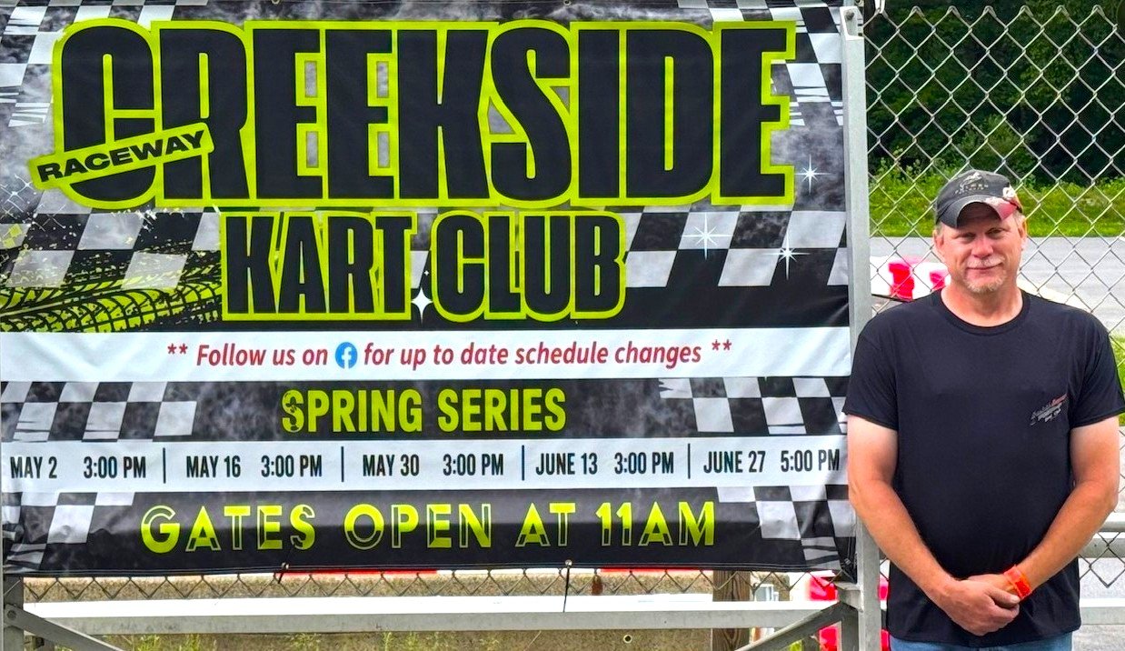 A man standing next to a large banner for Croekside Raceway Kart Club, displaying scheduled dates for spring racing events at the track, with the gates opening at 11 AM.