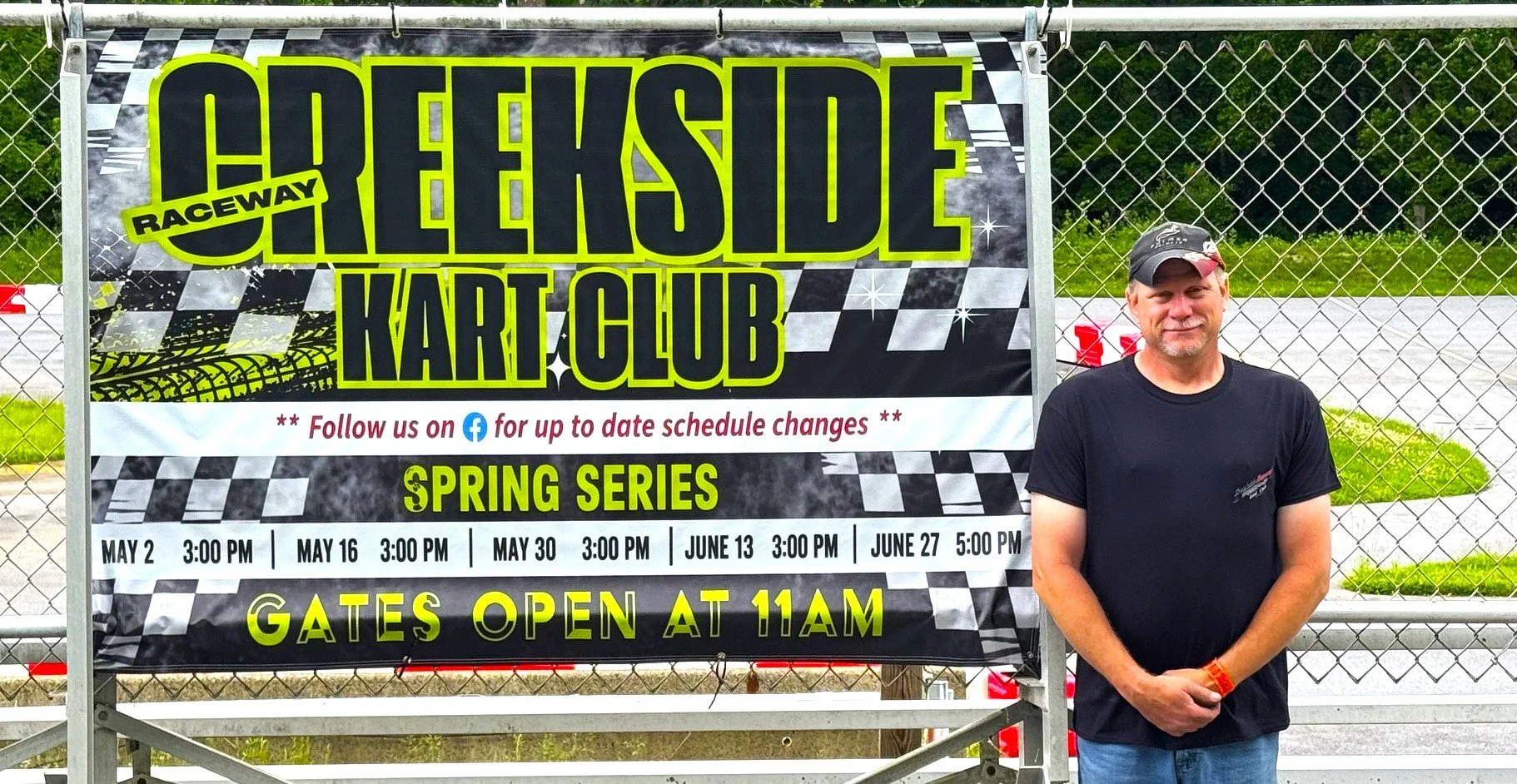 A man in a black t-shirt and cap standing next to a sign that reads 'Greenside Raceway Kart Club' on a chain-link fence, with race dates listed and gates opening at 11 AM.