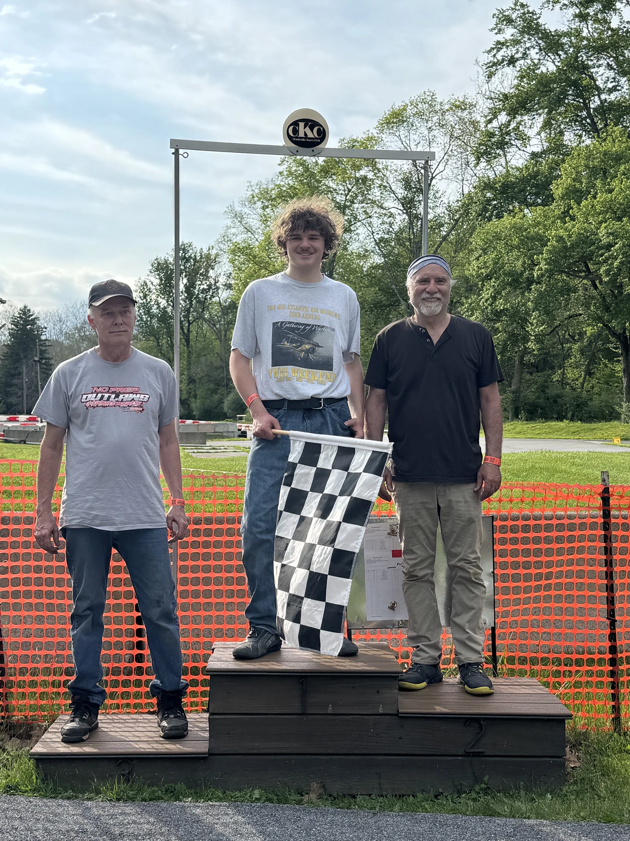 Three men standing on a podium with a checkered flag, outdoors, trees in the background, a banner with the logo "CKC" above them.