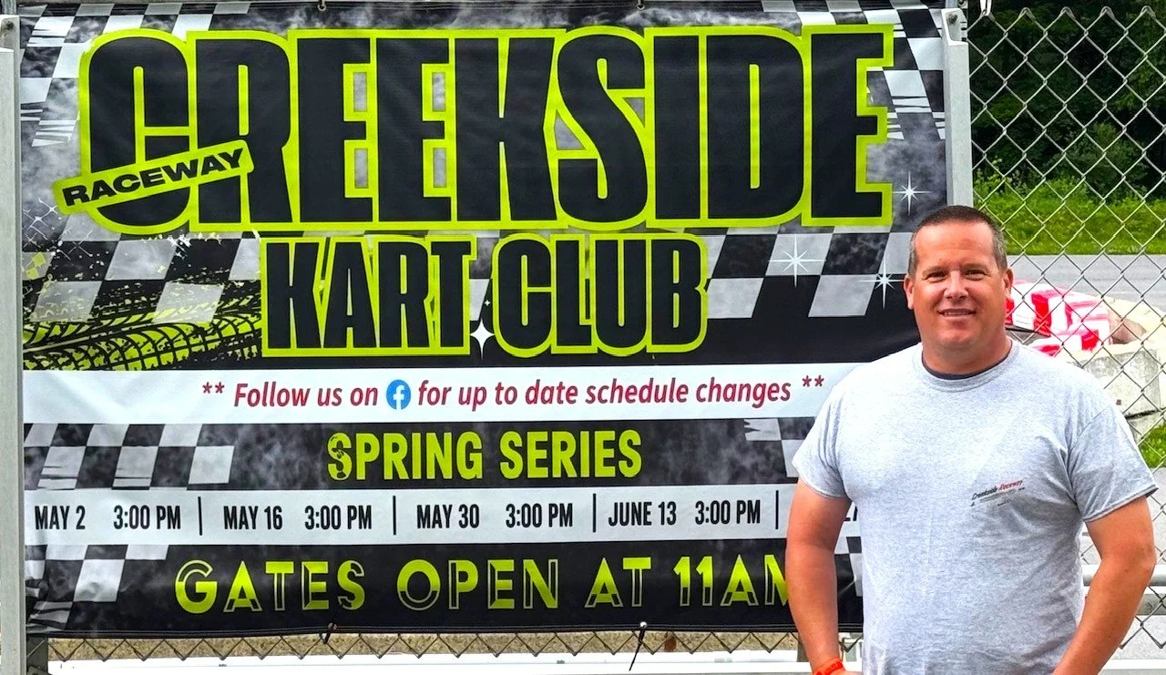 A man standing in front of a banner for Clearview Raceway Kart Club. The banner promotes the spring series with race dates listed and indicates that gates open at 11 AM. The man is wearing a gray t-shirt and smiling.