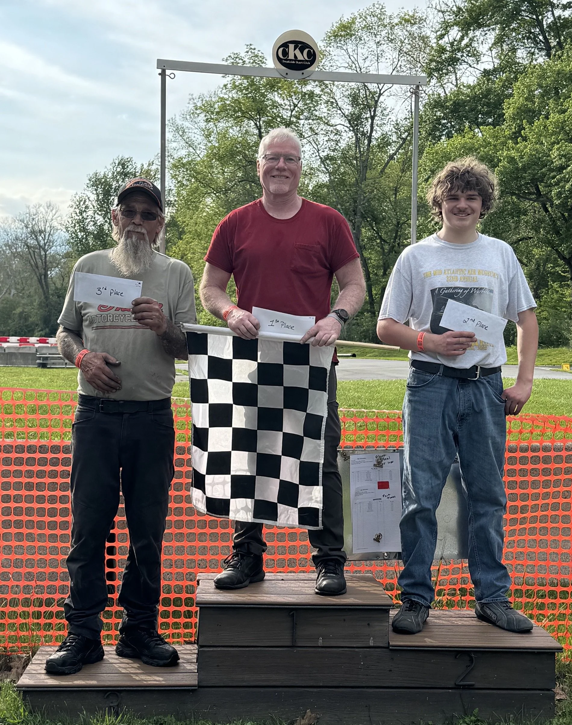 Three men standing on a winners' podium holding certificates at an outdoor event, with trees and a cloudy sky in the background.