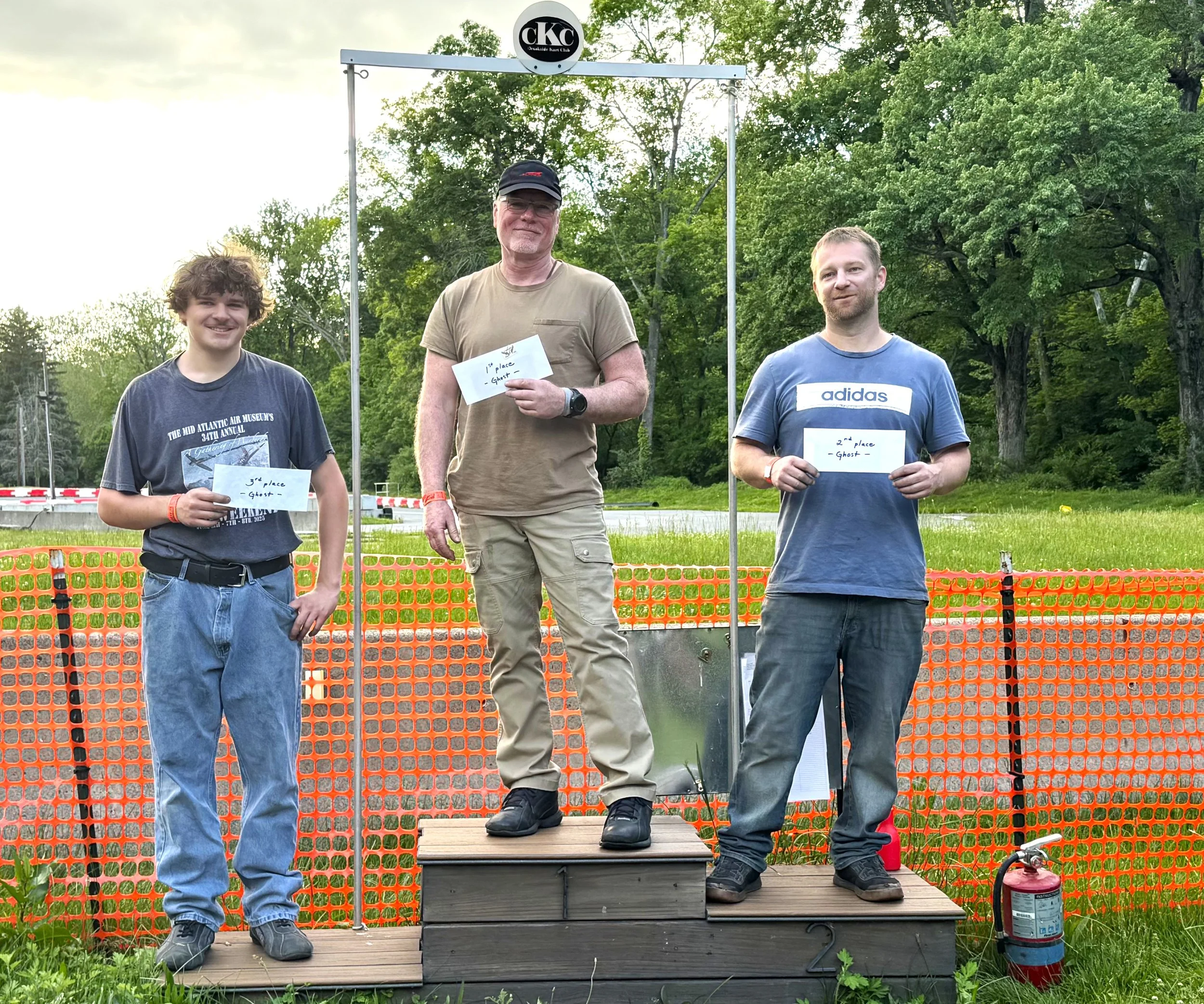 Three men standing on a wooden podium outdoors with a grassy field and trees in the background. The man in the center is on the highest level, holding a paper that says '1st place - Ghost', and is flanked by two men with papers reading '3rd place - Ghost' on the left and '2nd place - Ghost' on the right, all smiling. Behind them is a metal arch with a round sign reading 'CKC' and some orange safety fencing.