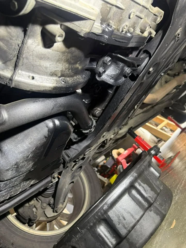 A close-up view of an automobile's undercarriage showing part of the engine, transmission, and suspension components in a garage setting.