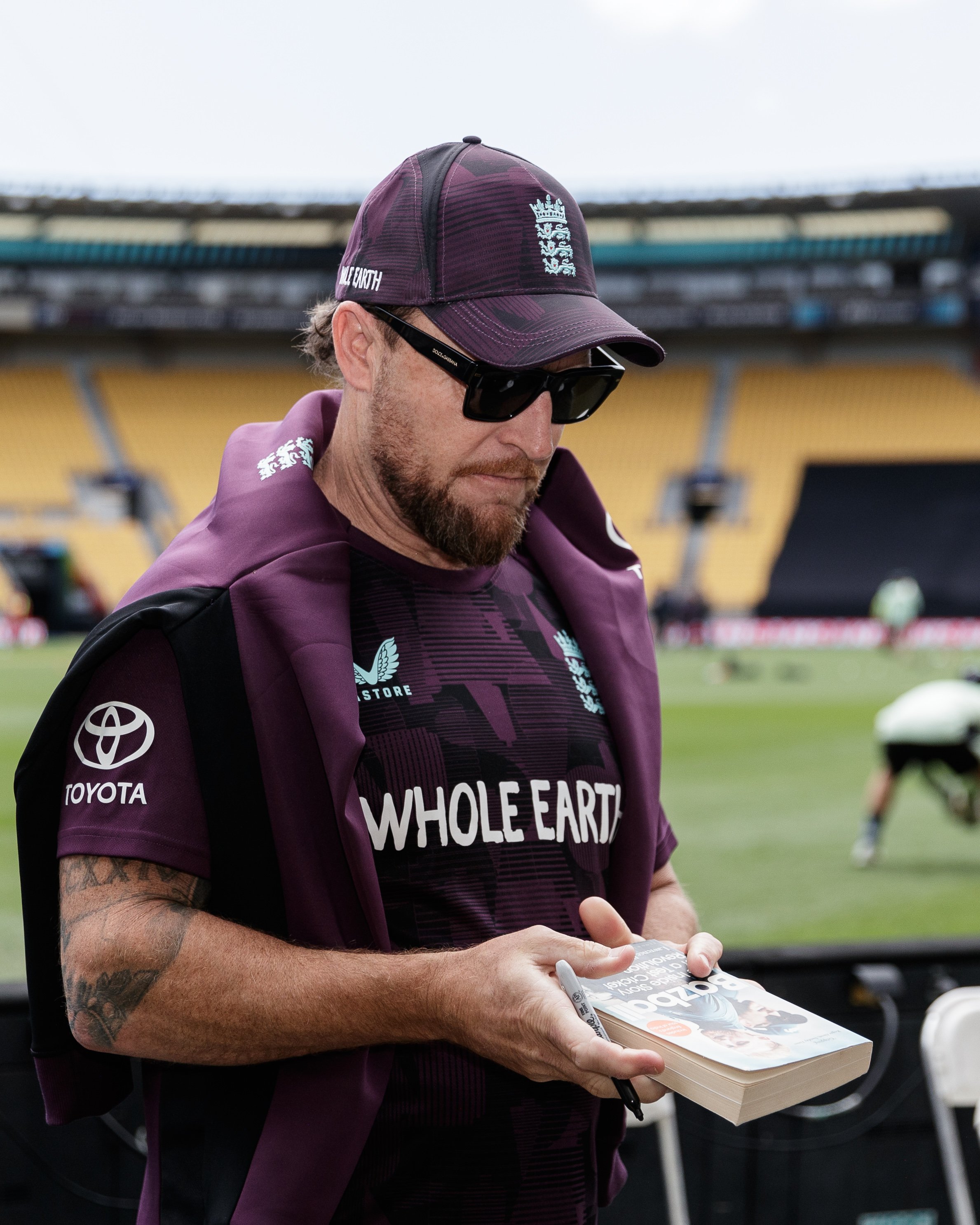 A man wearing sunglasses, a maroon cap with the England cricket logo, and a matching maroon shirt with sponsor logos, holding a newspaper and signing it at a sports stadium.