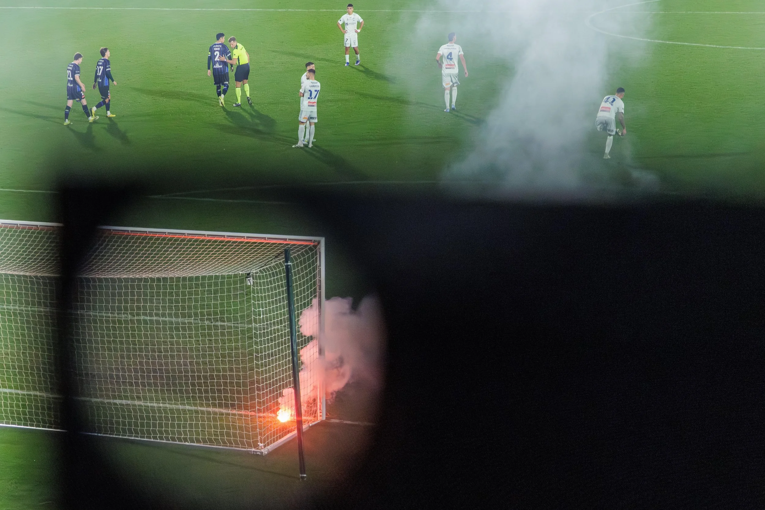 A soccer field with players and a referee near the center. Smoke or fog is visible, and a flare with bright orange flames and smoke is near the goal.