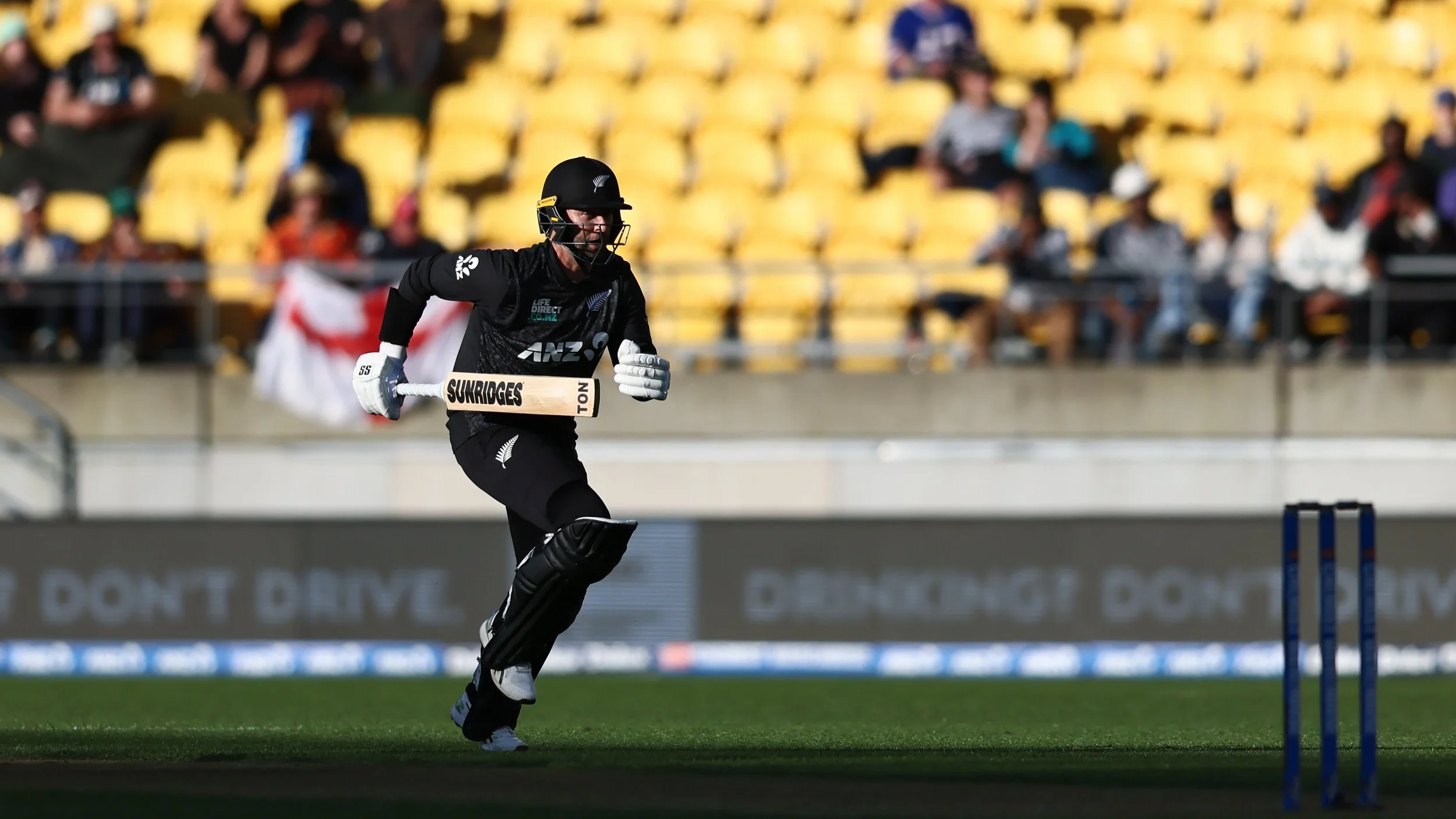 A cricket player in black uniform running on the field with a cricket bat in hand during a match, with yellow stadium seats and spectators in the background.