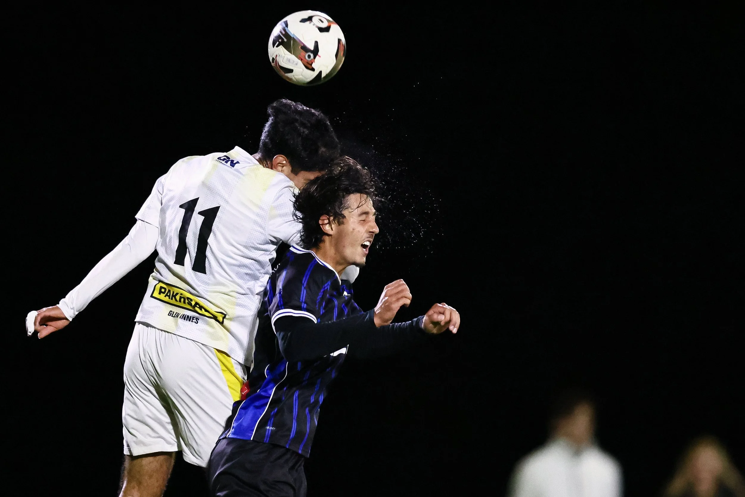 Two soccer players are jumping in the air competing to head a soccer ball during a night game. One player wears a white jersey with the number 11, and the other wears a dark jersey with blue stripes. The background is dark, emphasizing the action.