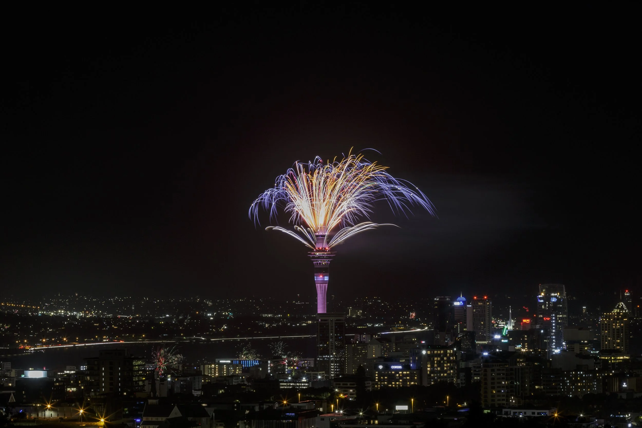 Nighttime cityscape with fireworks exploding from a tall tower.