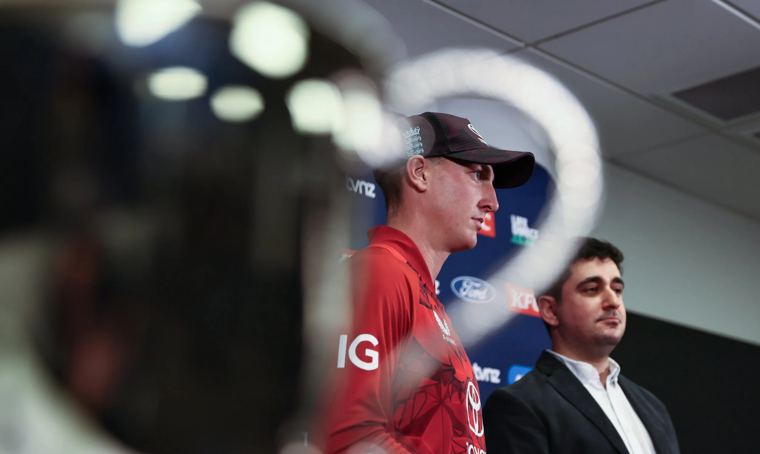 A man wearing a red sports shirt and a black cap with a logo, sitting at a press conference table, with two other men seated beside him, in front of a blue backdrop with sponsor logos.