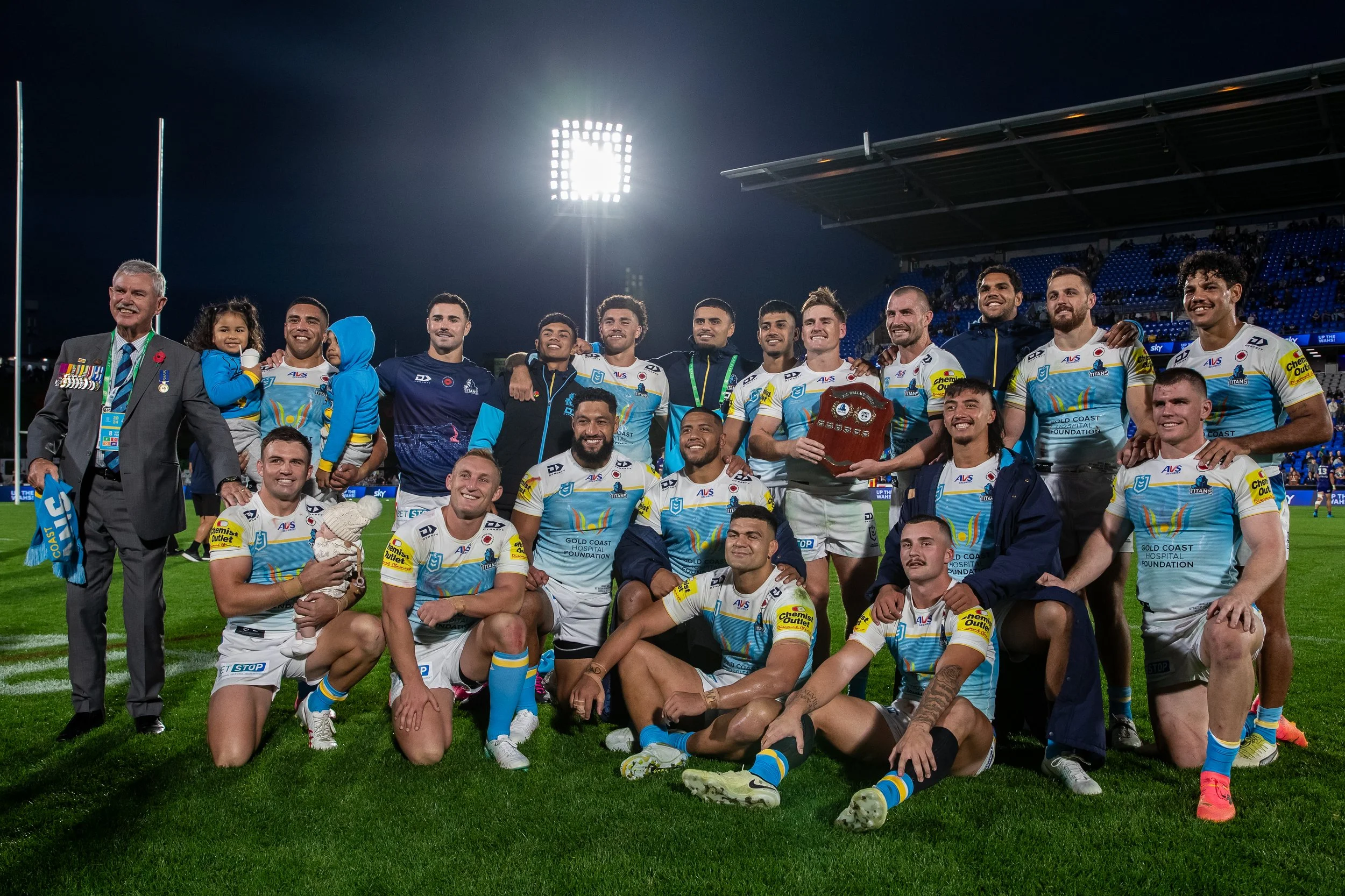 A rugby team celebrating on the field after a match, with players and staff smiling, holding a trophy, and posing for a group photo under stadium lights.