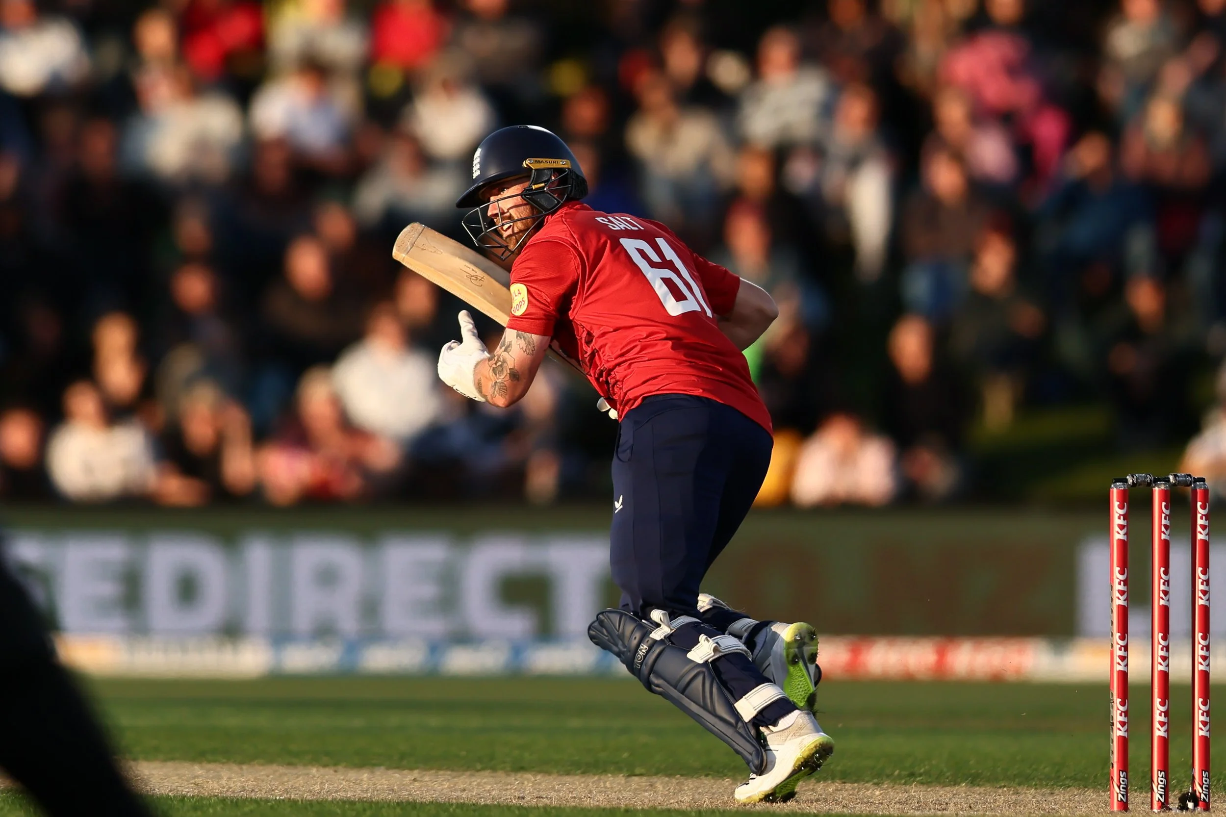 A cricket player wearing a red uniform and black helmet, hitting a ball with a wooden bat during a game in front of a blurred crowd.