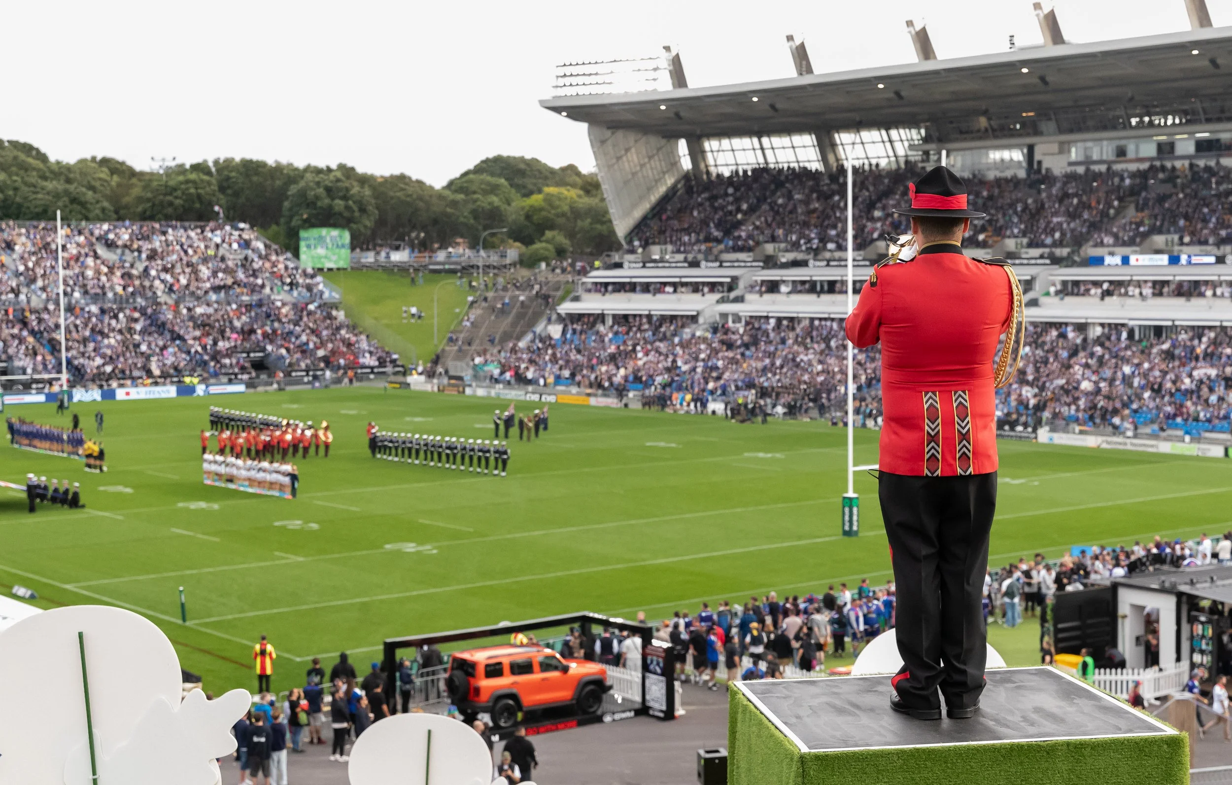 A marching band in red uniforms performing on a football field during a large outdoor event with a crowd in the stands and a modern stadium in the background.
